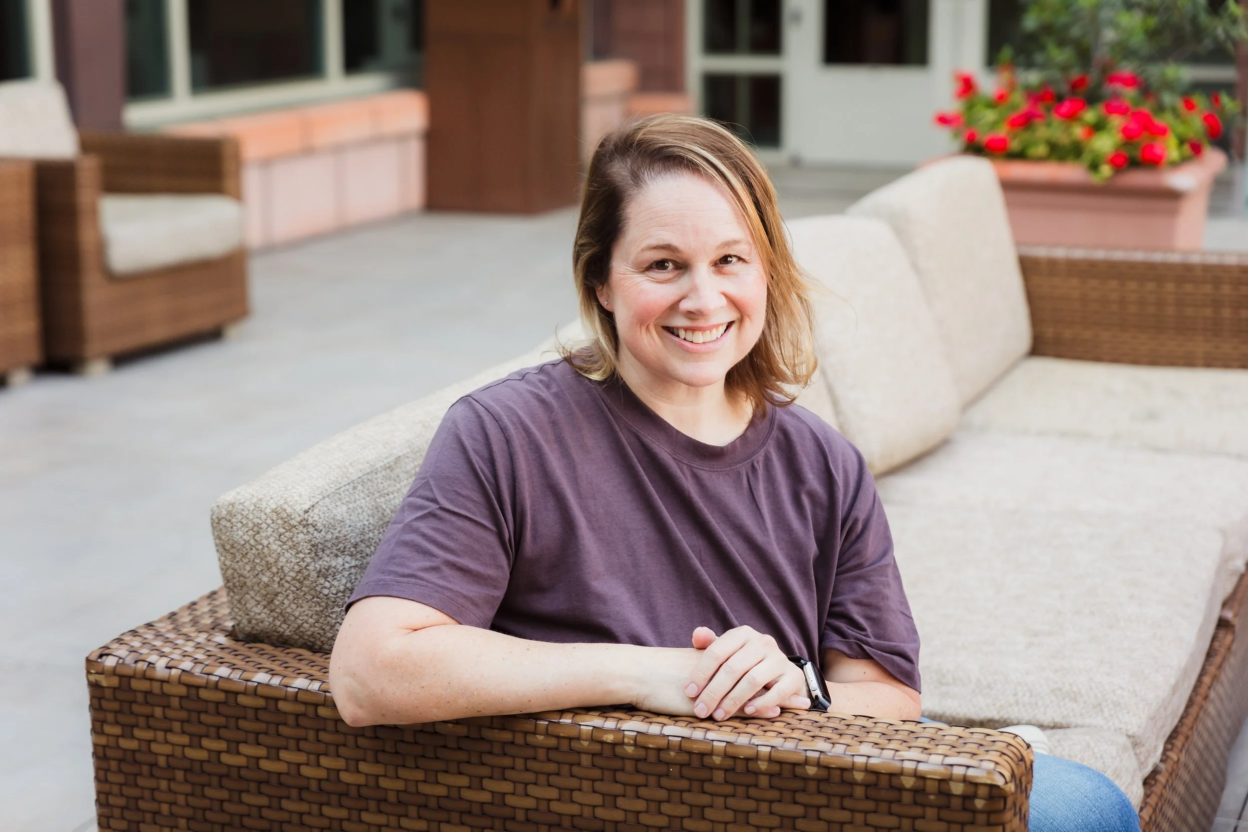 A woman with shoulder-length light brown hair, wearing a dark purple t-shirt,, sitting outside on a wicker chair, smiling at the camera. In the background, there are brick walls, large windows, and a large planter with bright red flowers.