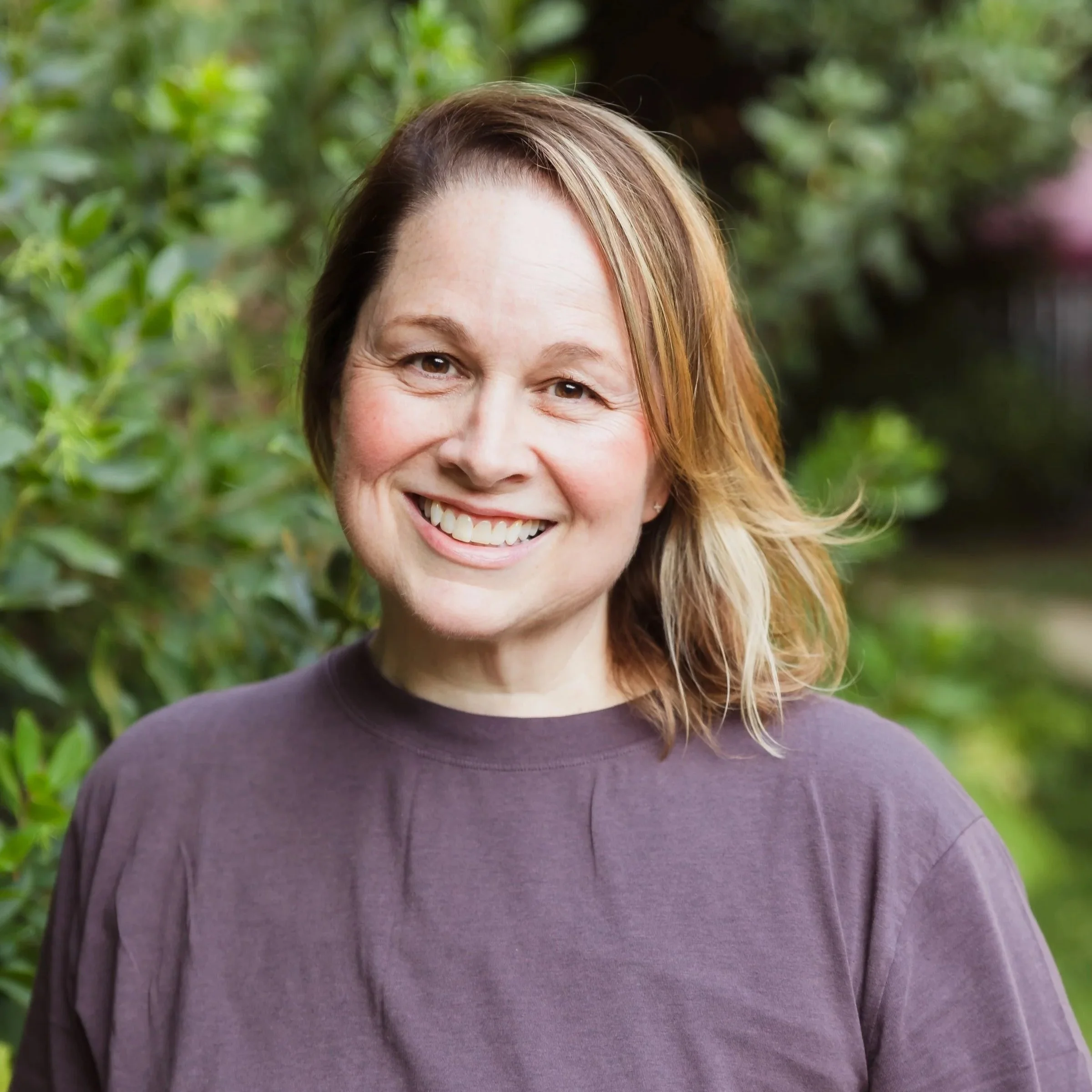 Close-up of a smiling woman with light brown, slightly wavy hair, wearing a purple shirt, outdoors with green foliage in the background.