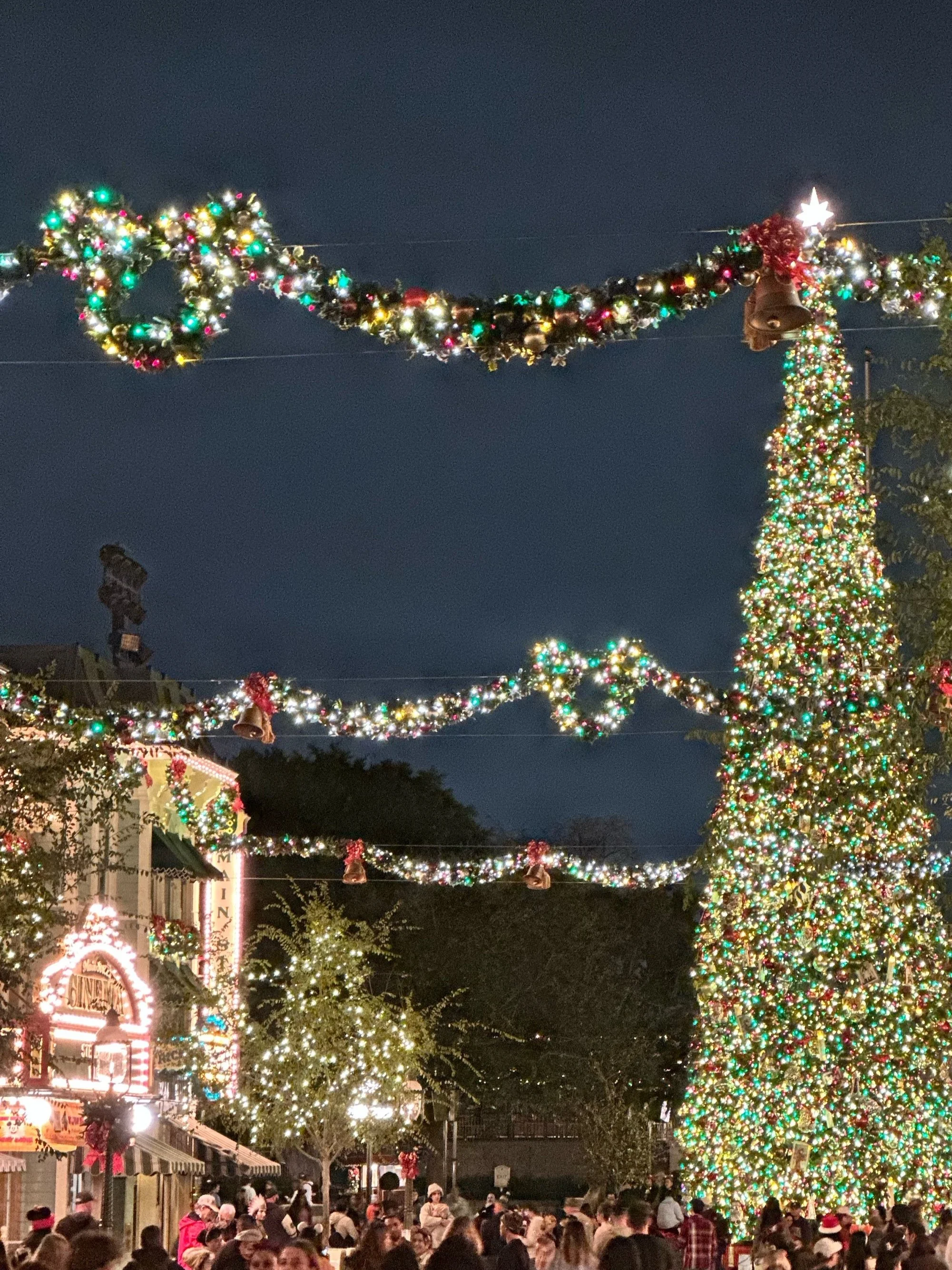 Festive street scene at night with a large Christmas tree adorned with colorful lights, garlands, and decorations. Illuminated buildings and holiday lights create a cheerful atmosphere. A crowd of people is visible, enjoying the festive setting.