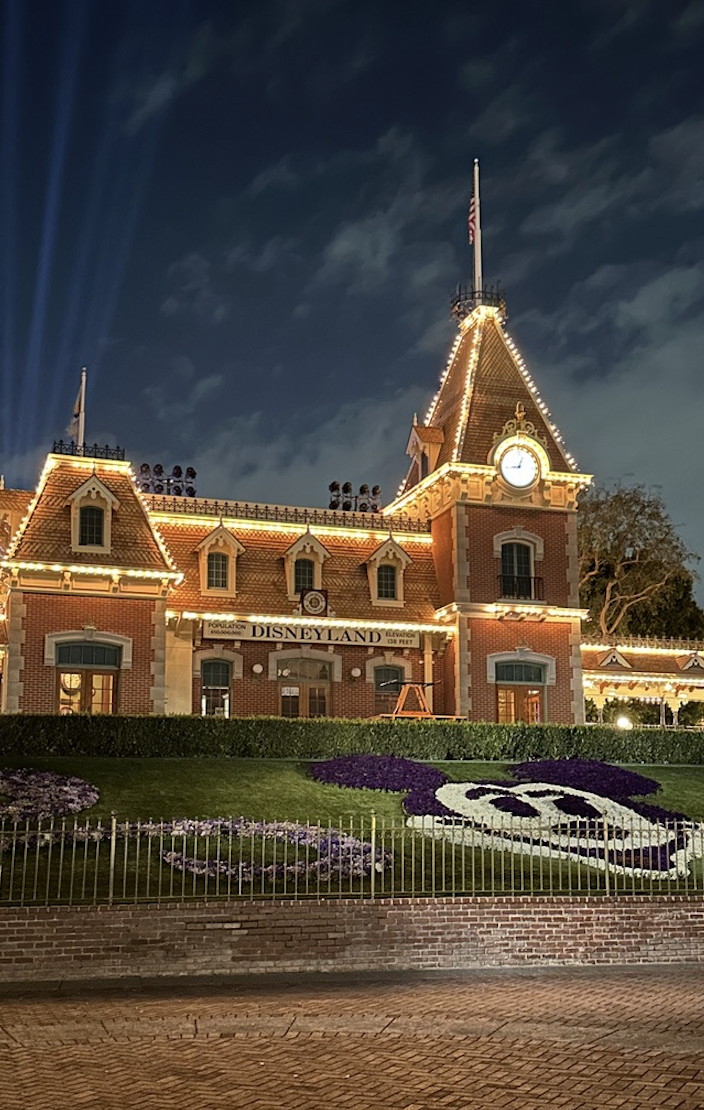 Disneyland entrance at night, illuminated with lights, featuring a clock tower and Mickey Mouse floral display.