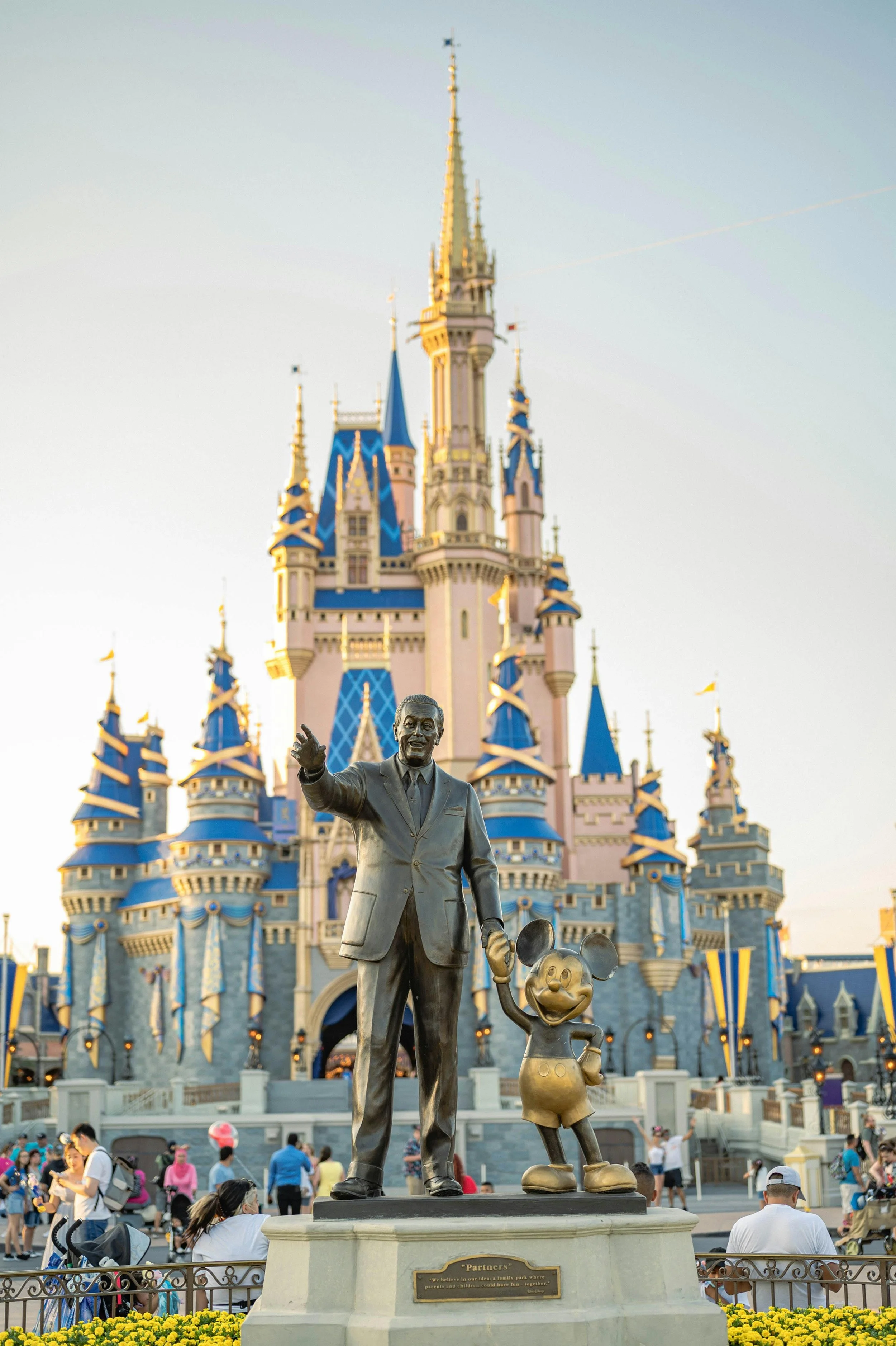 Statue of a man and Mickey Mouse in front of a castle at a theme park with visitors.