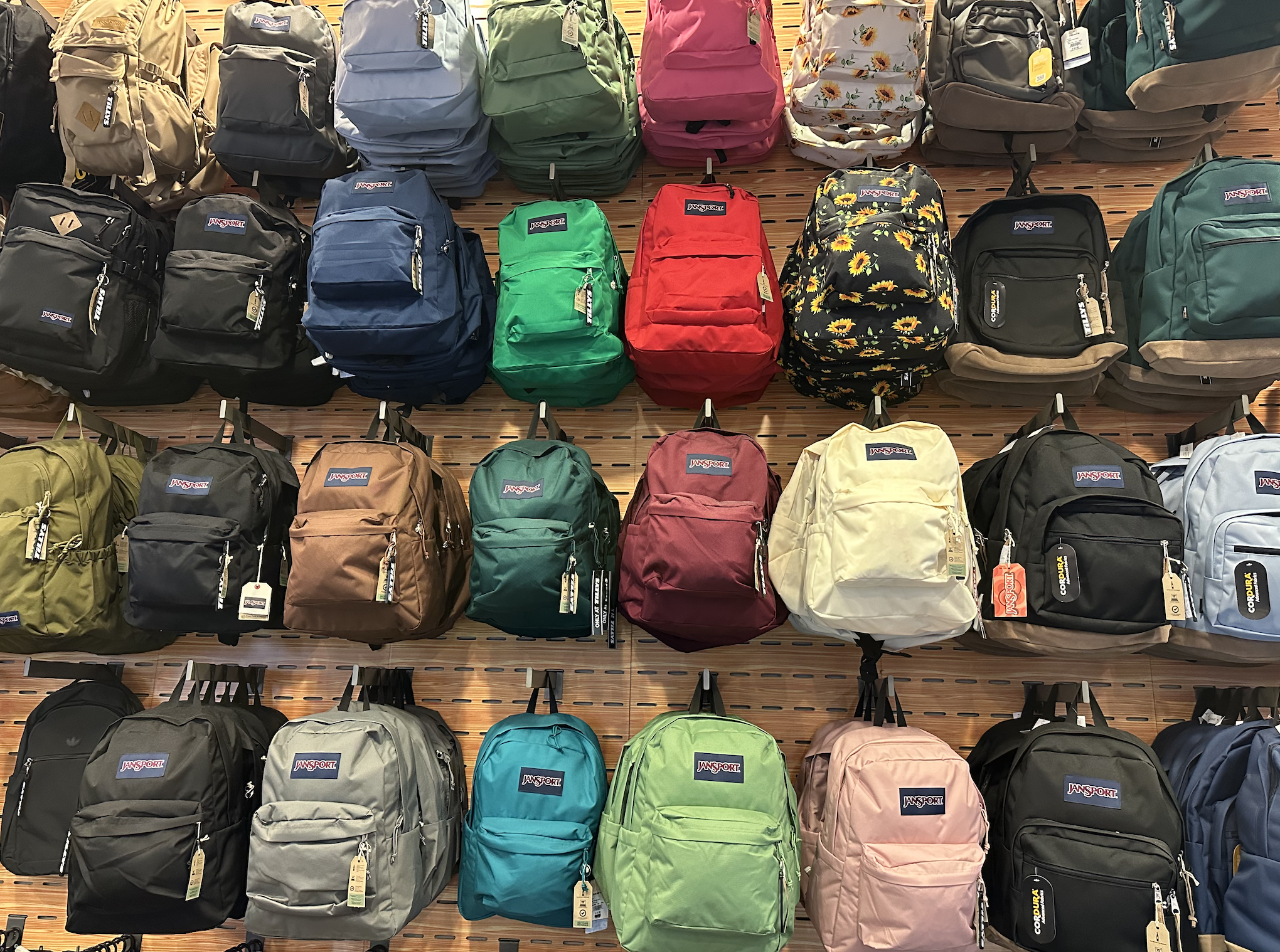 a-wall-of-multi-colored-jansport-backpacks-hanging-on-hangers-on-a-wood-paneled-wall