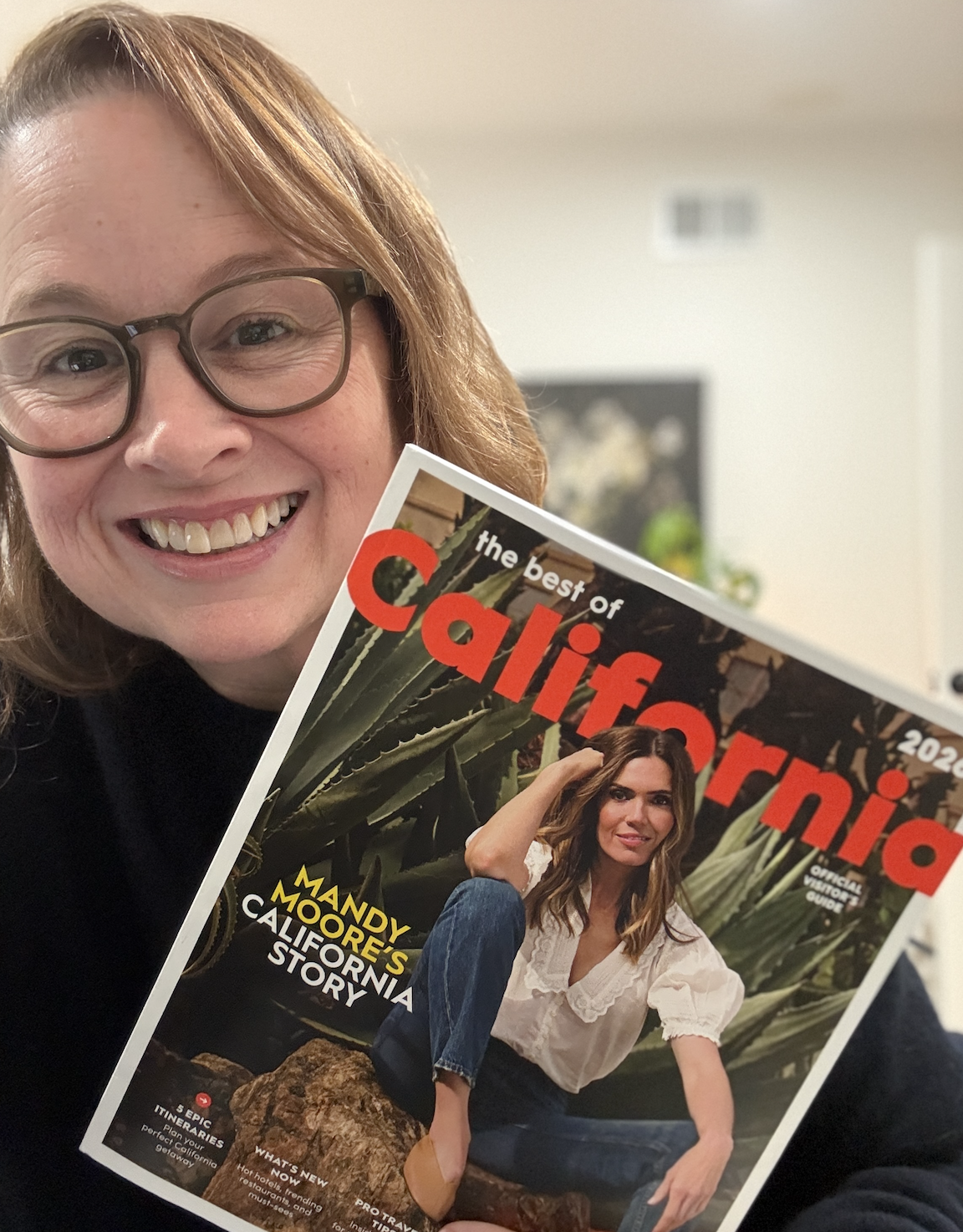 A woman with glasses smiling and holding a magazine titled 'The Best of California' featuring a woman in a white blouse and jeans sitting among large agave plants.