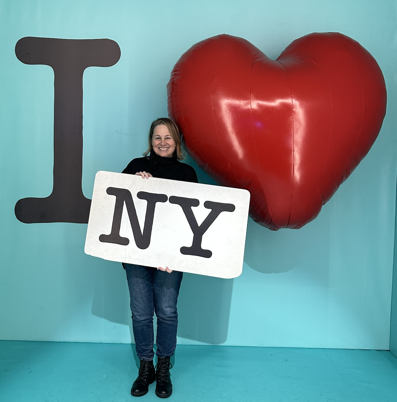 A woman smiling while holding a large sign that says 'NY' in front of a red heart-shaped balloon, with a background featuring a large 'I' and the balloon.