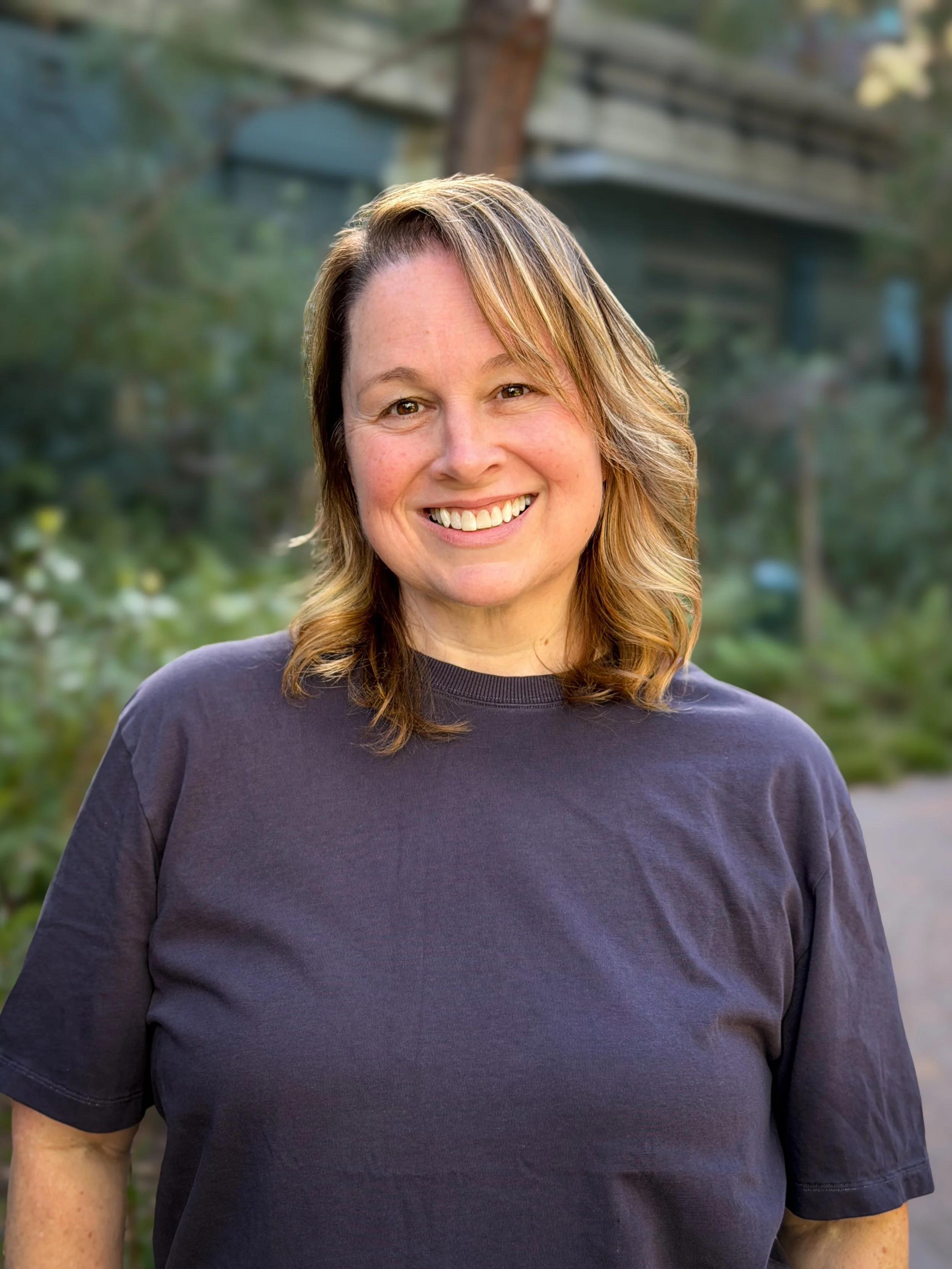 Close-up of a middle-aged woman with shoulder-length light brown hair with blonde highlights, smiling, outdoors in front of a blurred background of trees and a house.