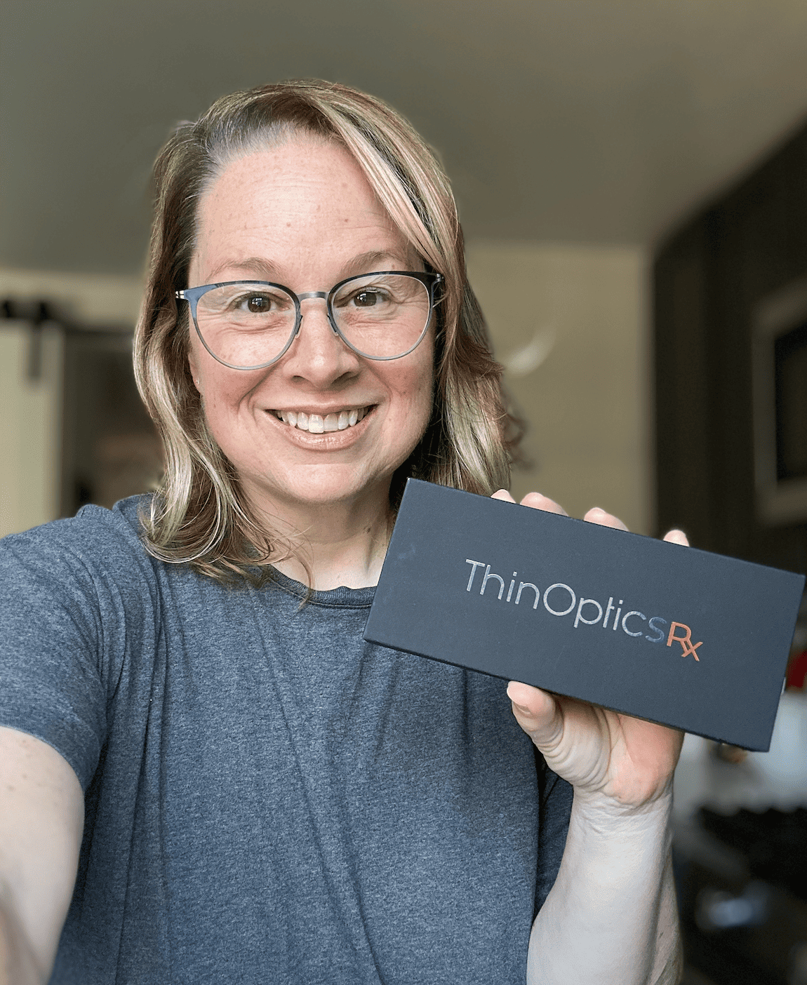 Woman smiling and holding a box labeled 'ThinOptics RX' in a home setting.