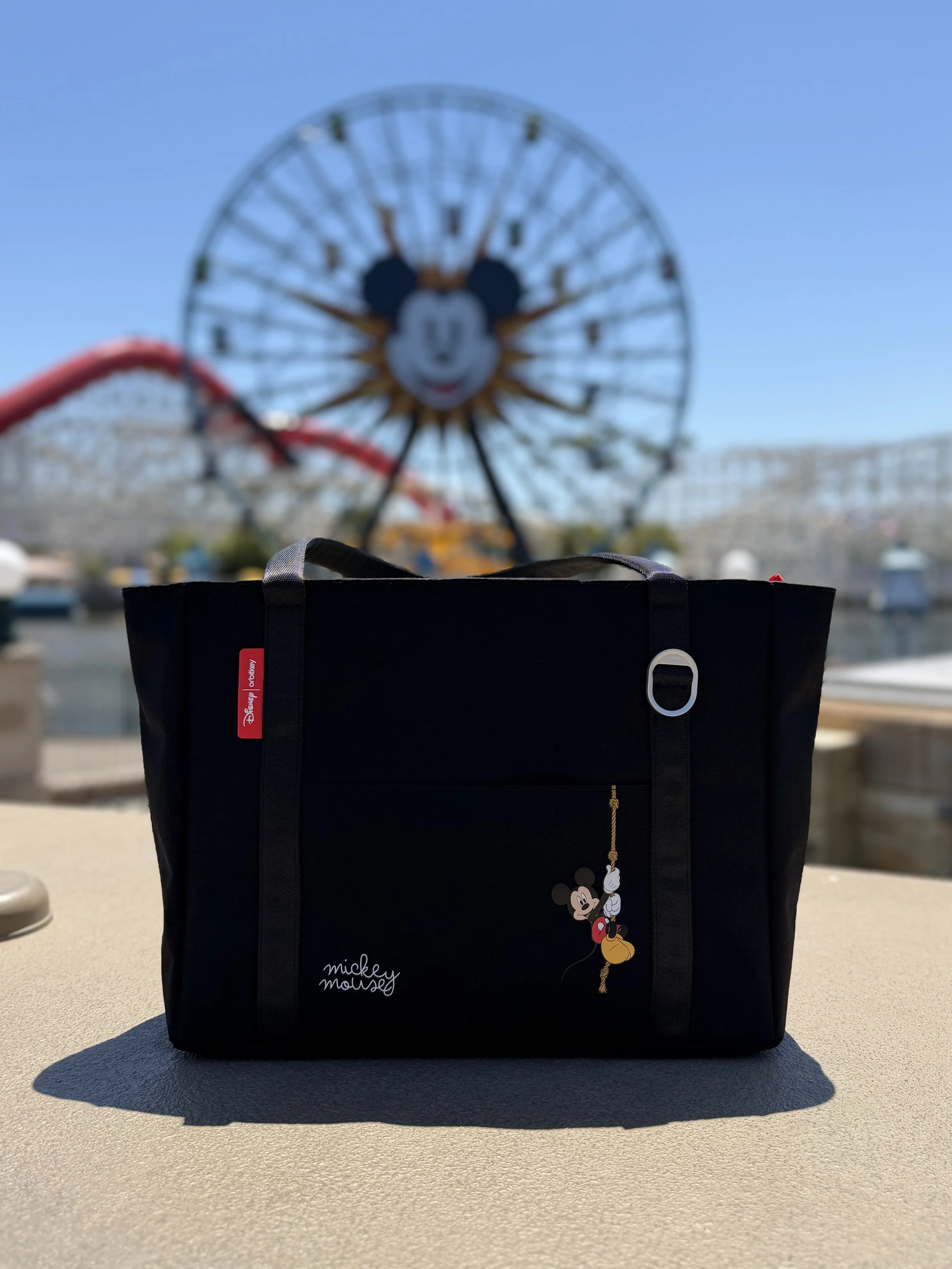 A black bag with a Mickey Mouse design and the words "mickey mouse" hanging from a hook, placed on a table outdoors with a Disney theme park Ferris wheel featuring Mickey Mouse's face in the background.