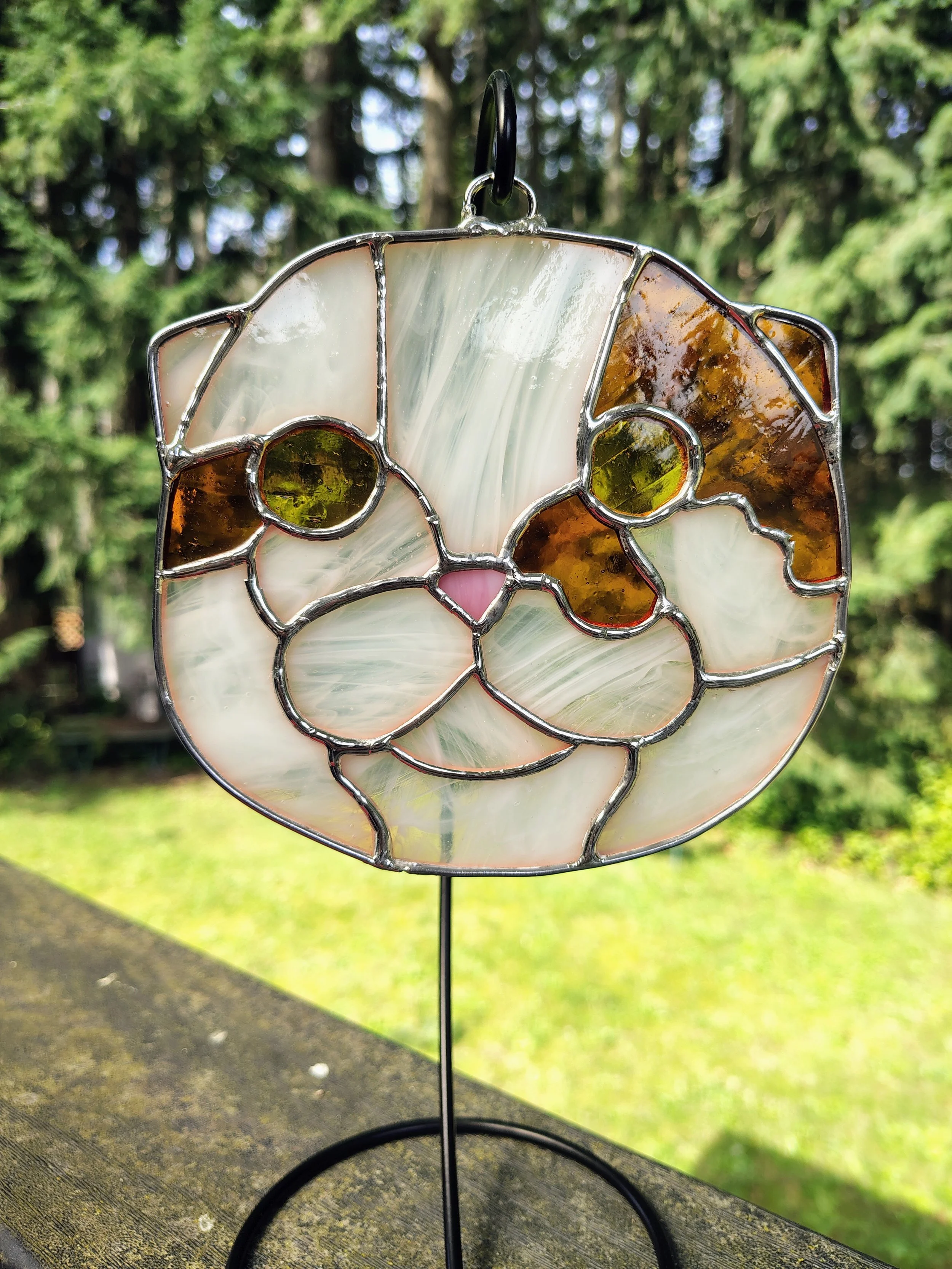 an orange and white cat made of stained glass on a black hook in front of trees and grass