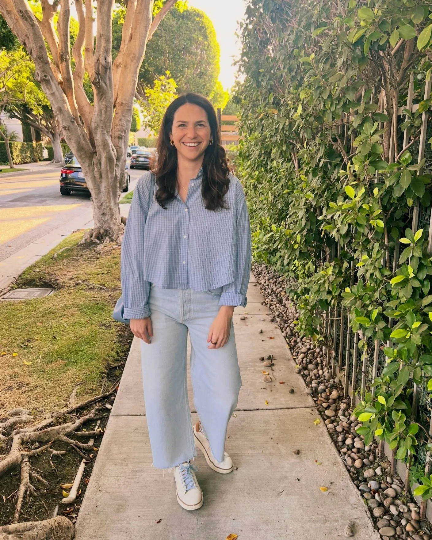 A woman with dark hair, wearing a blue checkered shirt, light blue wide-leg jeans, and white sneakers, smiling and standing on a sidewalk surrounded by trees and greenery in a sunny outdoor setting.