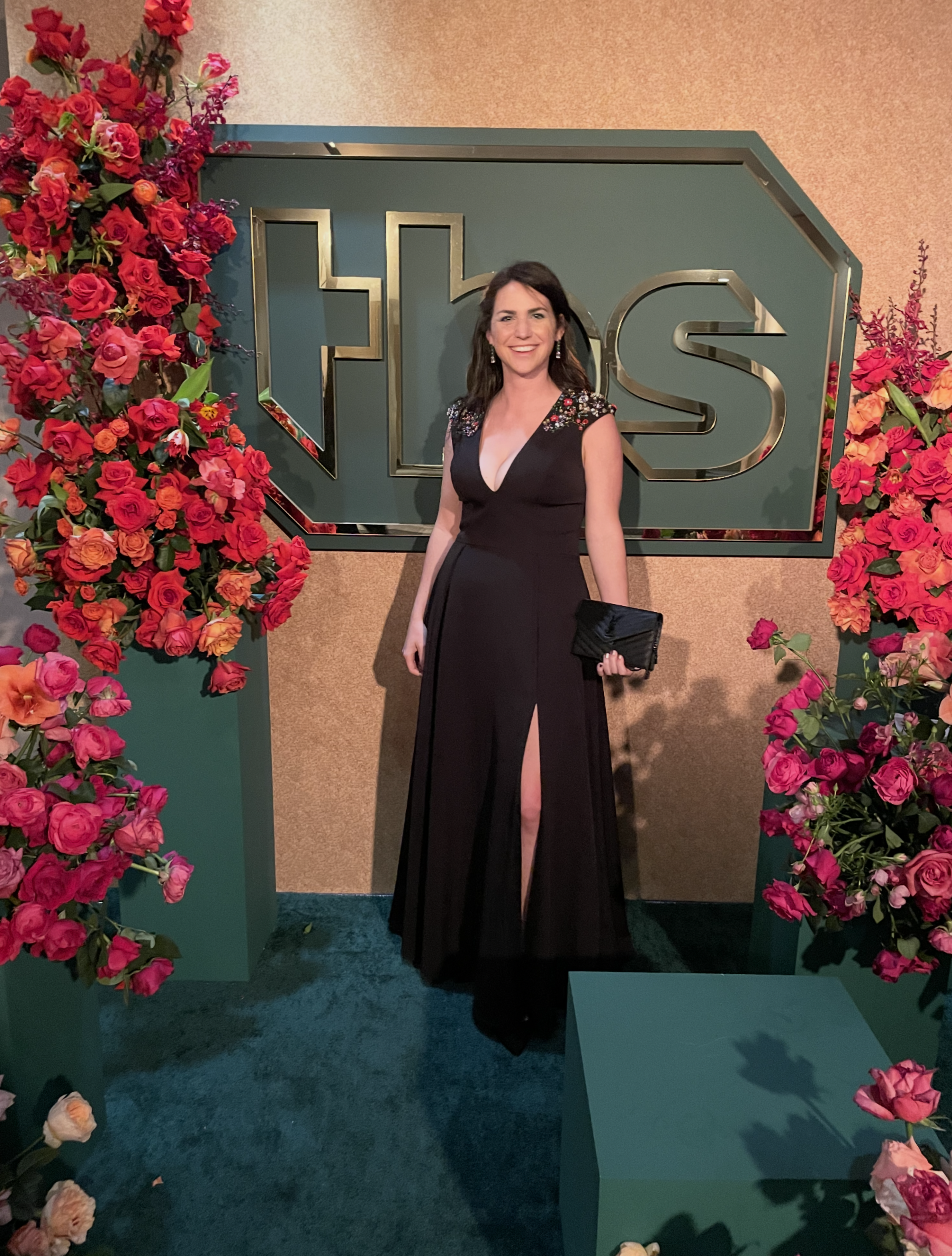 Woman in a black gown with floral embellishments on the shoulders, holding a clutch, standing in front of a backdrop with the word 'FLEA' in metallic letters, surrounded by pink and orange flowers.