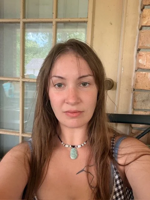 A woman with long brown hair takes a selfie in front of a window and brick wall, wearing a necklace and a sleeveless top.