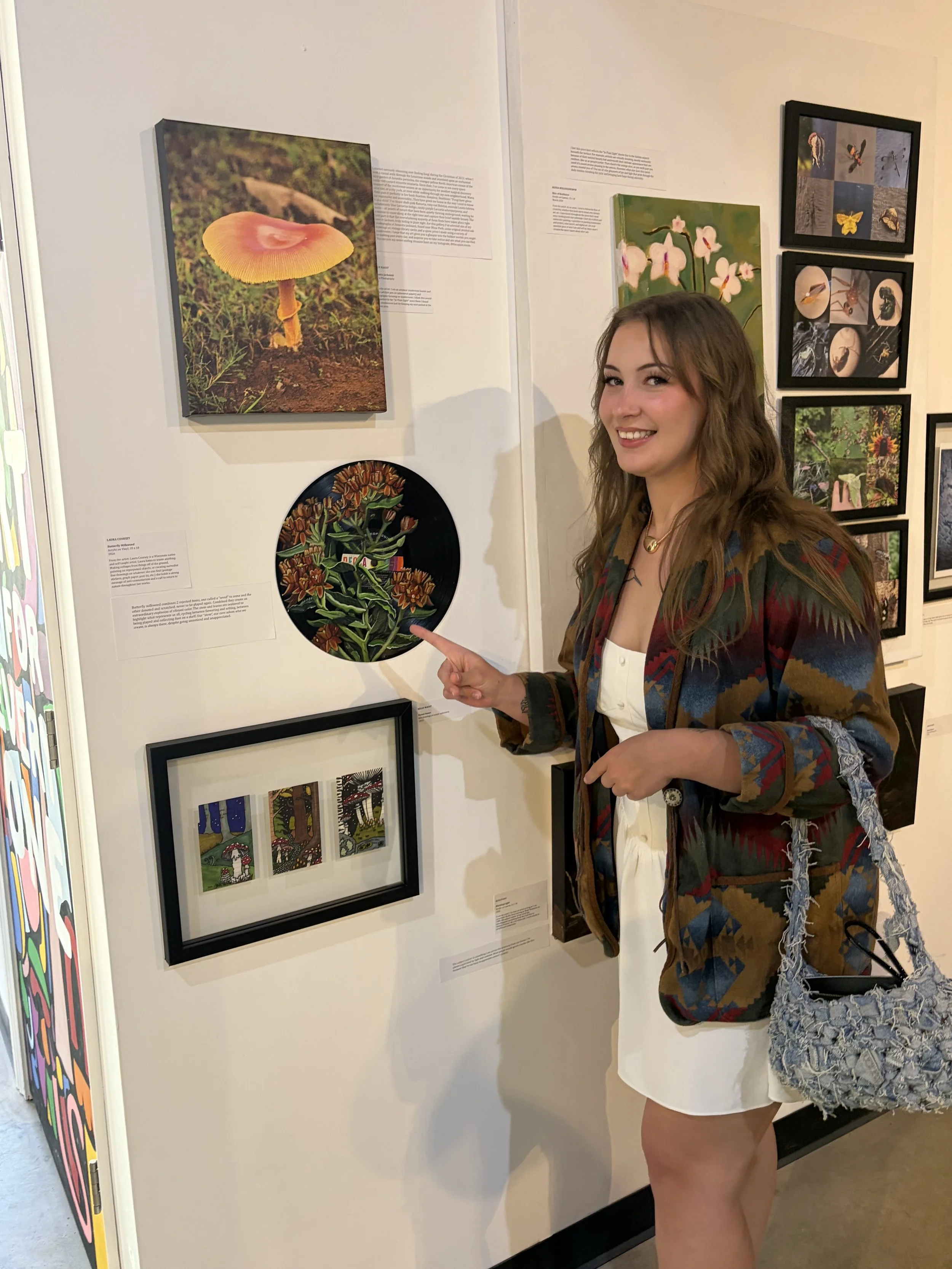 A woman at an art gallery pointing at a framed painting of orange and red flowers against a green background.
