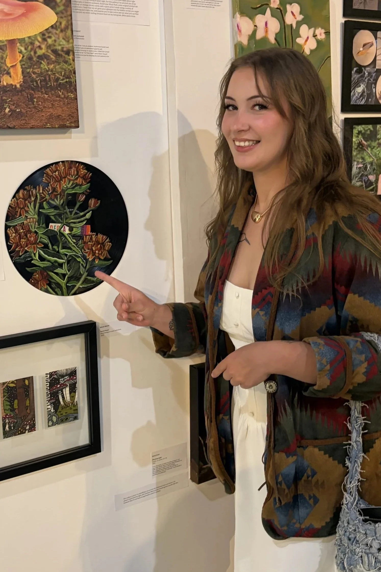 A smiling woman with wavy brown hair pointing at a framed artwork, at an art exhibition, with other framed artworks on the wall behind her.