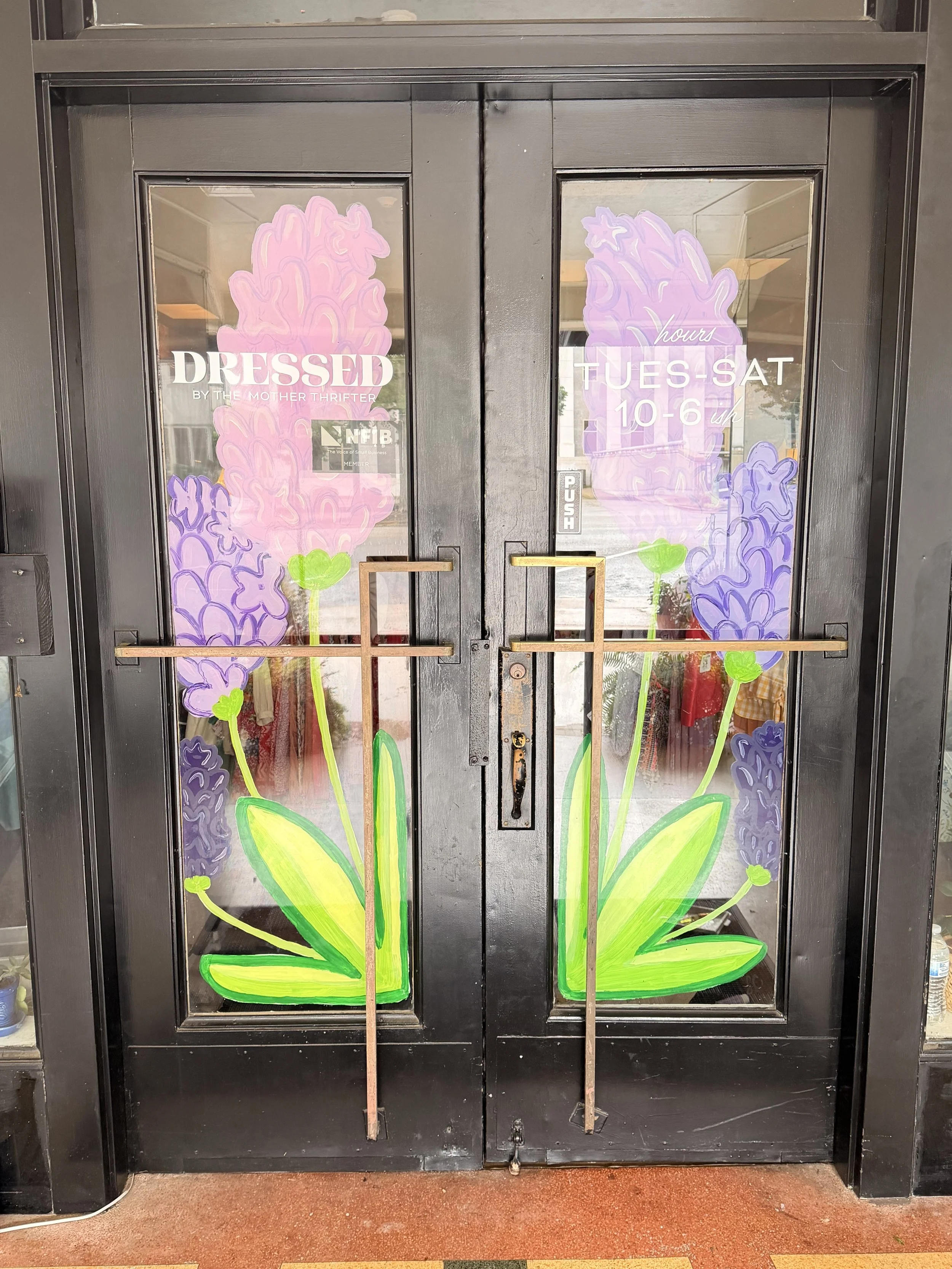 Decorative floral design on glass door with purple flowers, green leaves, and pink accents, including business hours and sign for dress shop.