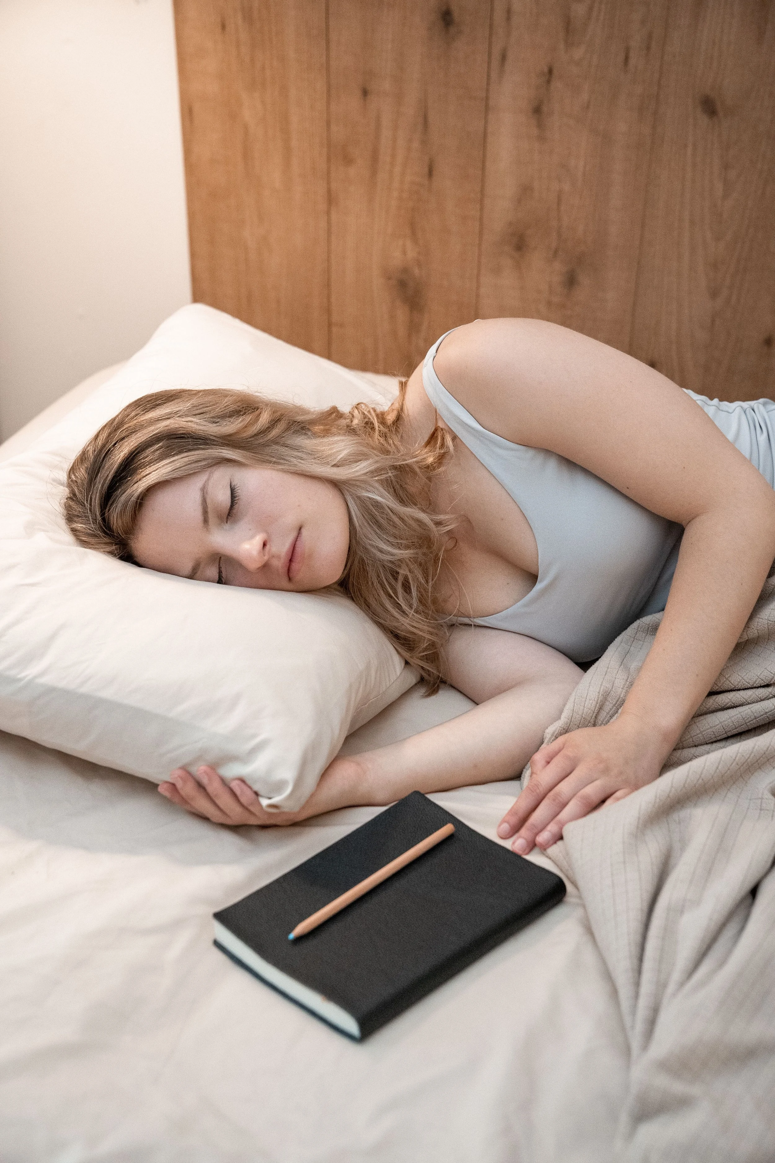 A young woman sleeping on a bed with a white pillow, wearing a sleeveless light blue top. A black notebook with a brown pencil rests on the bed near her. The background features a wooden headboard and a white wall.