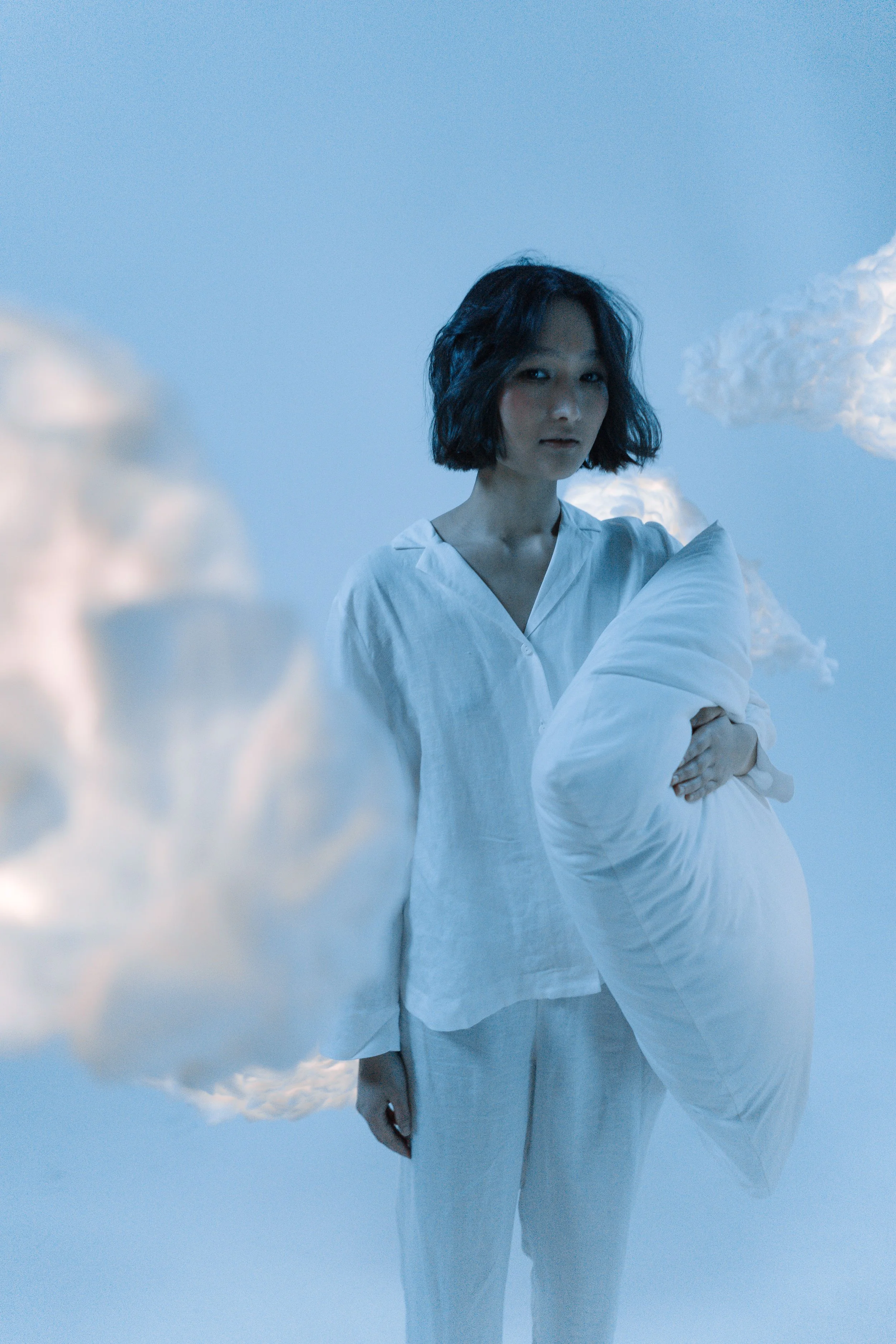 A woman with short black hair wears a white shirt and white pants, standing against a blue sky with clouds, holding a white pillow, with clouds in the background.