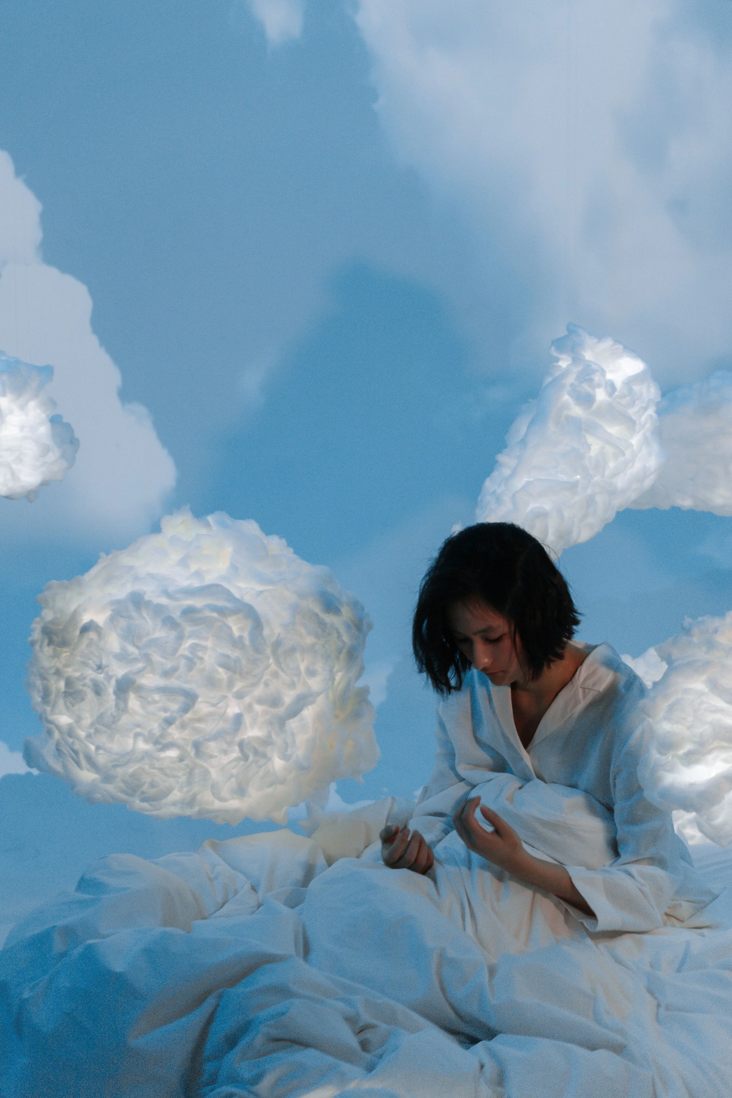 A woman with dark hair and light skin is sitting on a floor covered with white fabric, surrounded by cloud-shaped light fixtures hanging from a blue sky backdrop, creating an artistic scene.