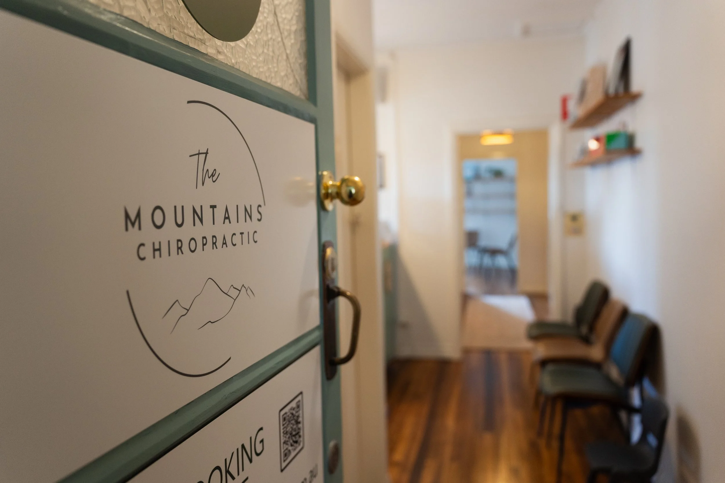 Close-up of a sign on a wall that reads 'The Mountains Chiropractic' with a mountain outline below. The background shows a hallway with chairs and shelves.