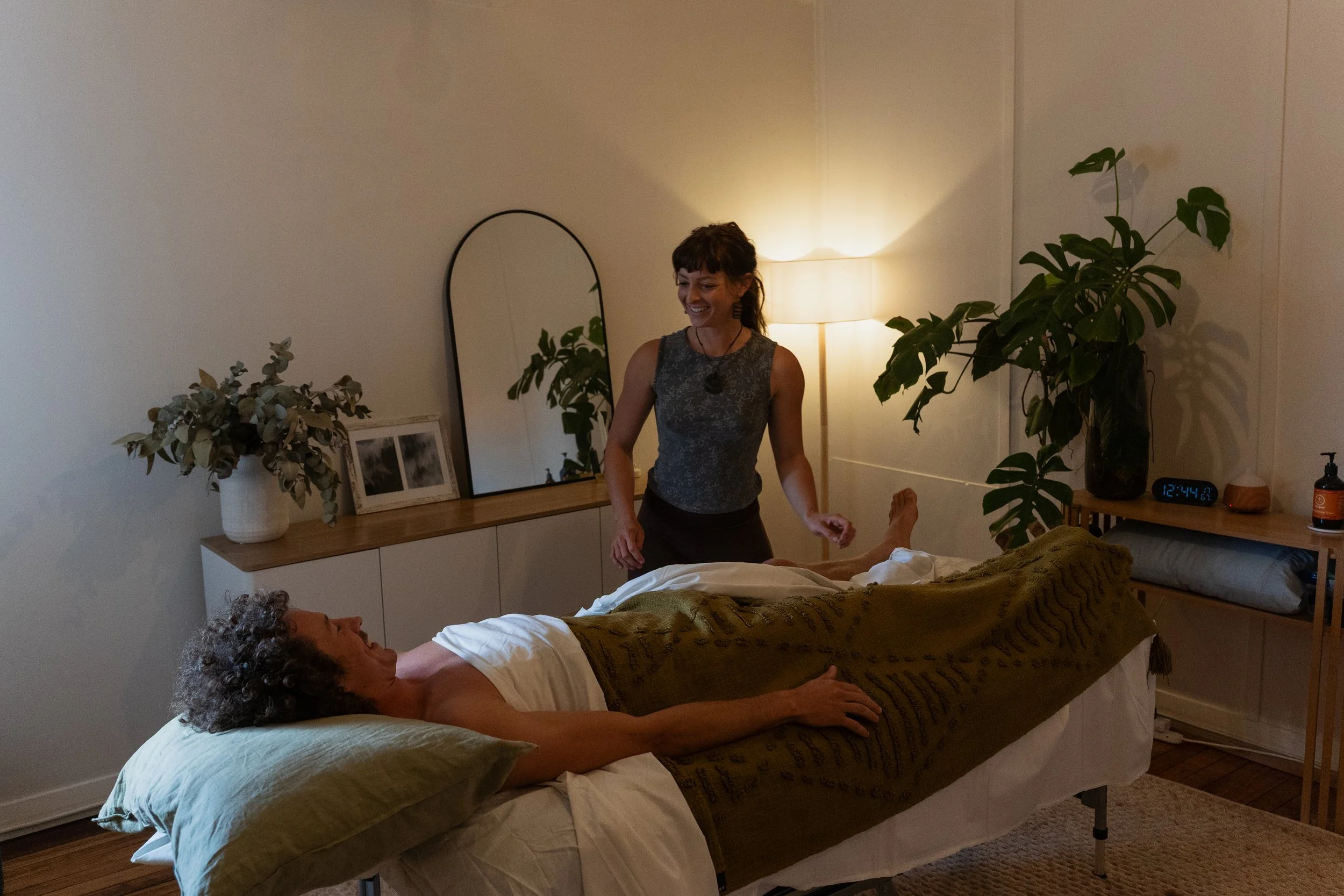 A woman with curly gray hair lying in bed, covered with a brown blanket, and another woman with dark hair smiling and standing beside the bed in a bedroom with a large plant, a mirror, and a lamp