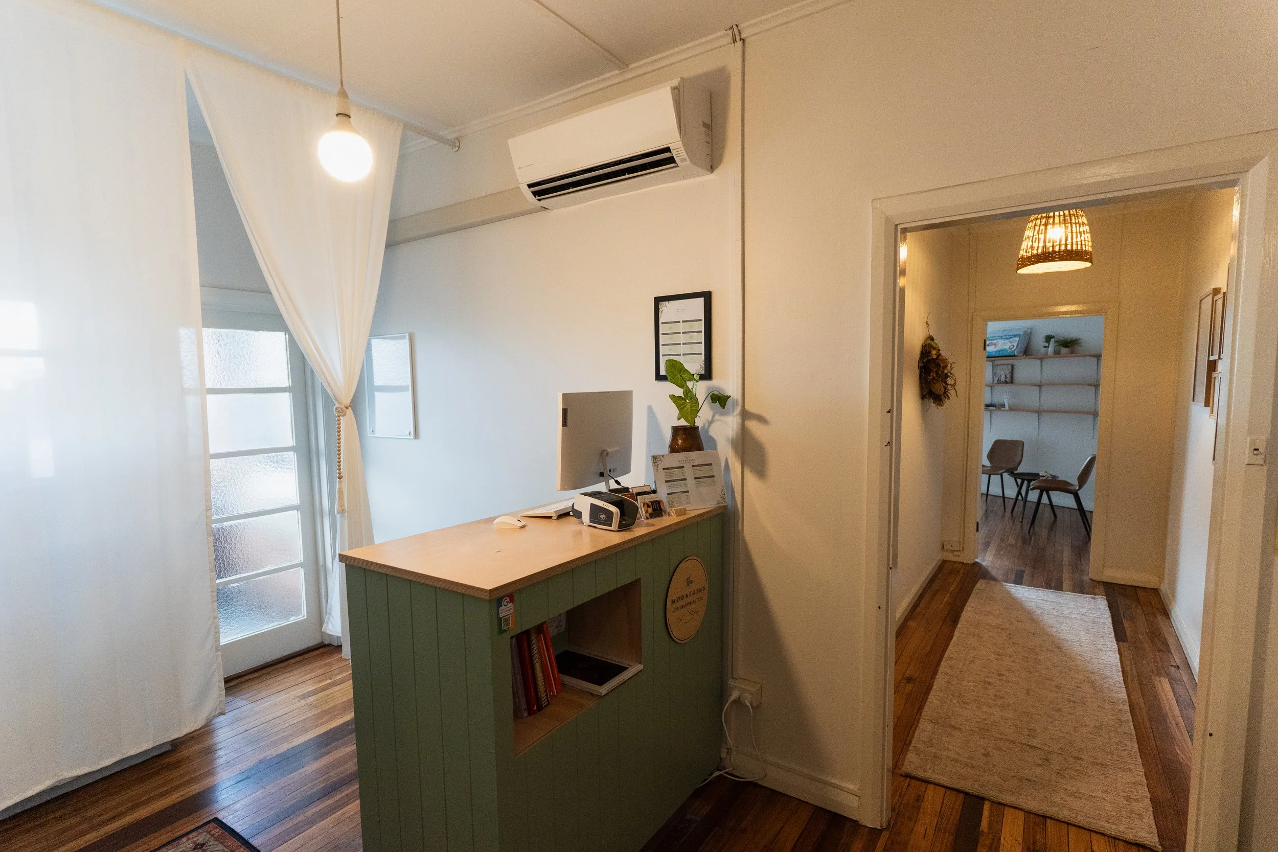 Interior of a cozy home with a reception desk, air conditioning unit on the wall, and a doorway leading to a small seating area with chairs and a bookshelf.