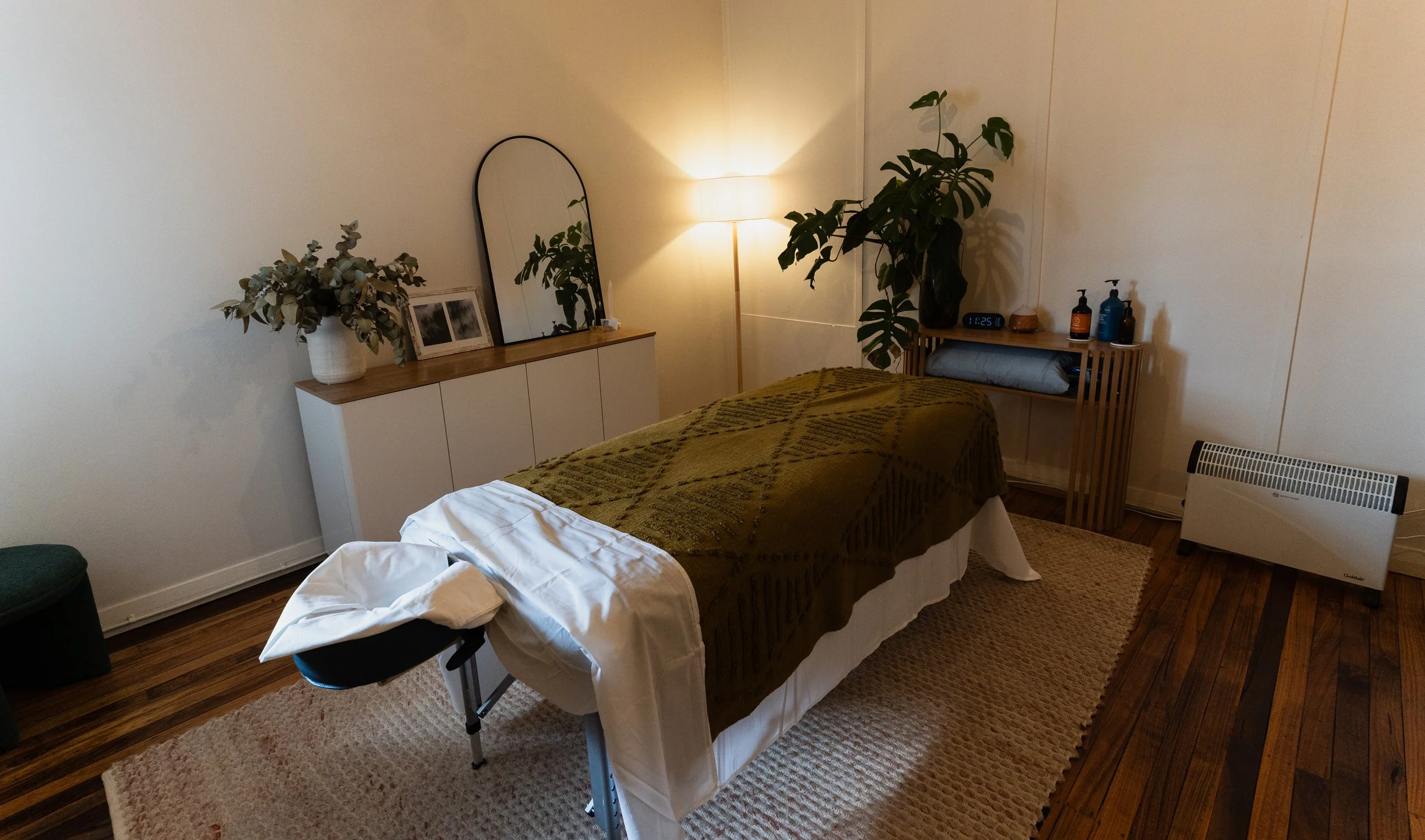A massage or treatment room with a massage bed covered with a green blanket, a pillow, and a small stool nearby. There is a white cabinet with decorative items, a mirror, and a lamp. Potted plants are in the corner, and a digital clock is on a wooden