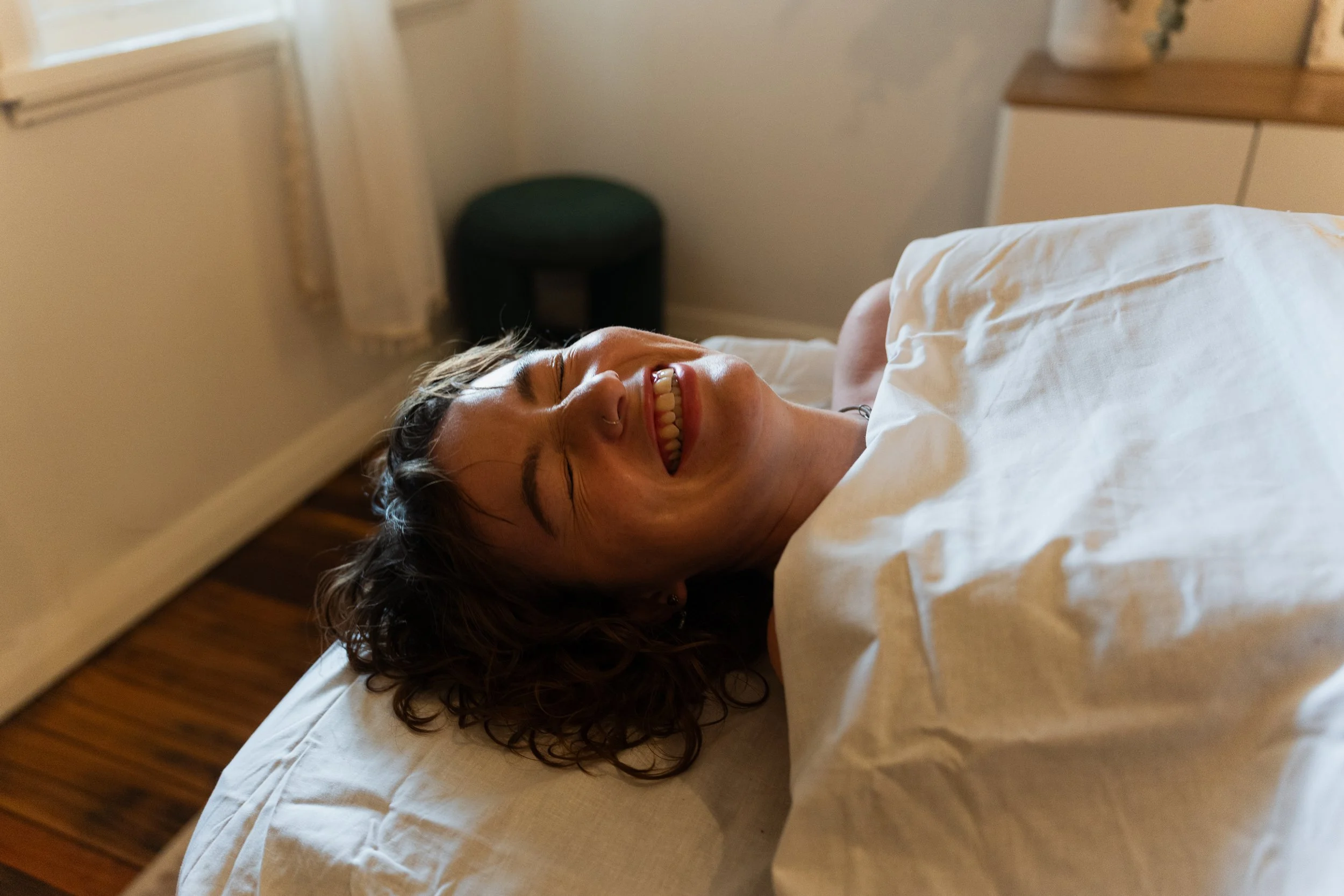 Person with curly hair smiling and lying on a bed, with beige walls, a window with curtains, a nightstand, and a plant in the background.