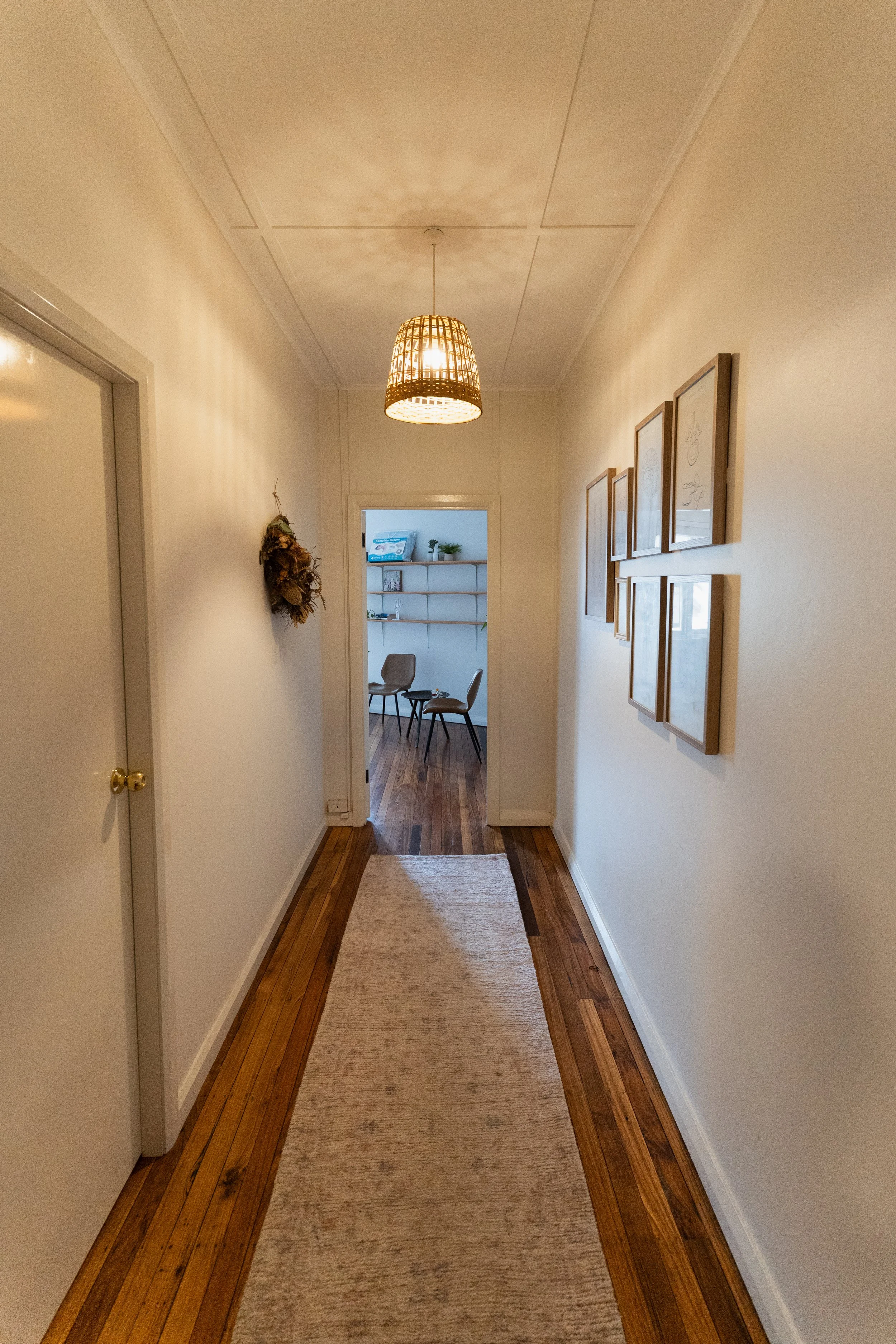 A hallway with white walls, wooden floor, hanging light fixture, framed artwork on the right wall, a doorway on the left leading to a room with chairs and a shelf, a beige runner rug on the floor.