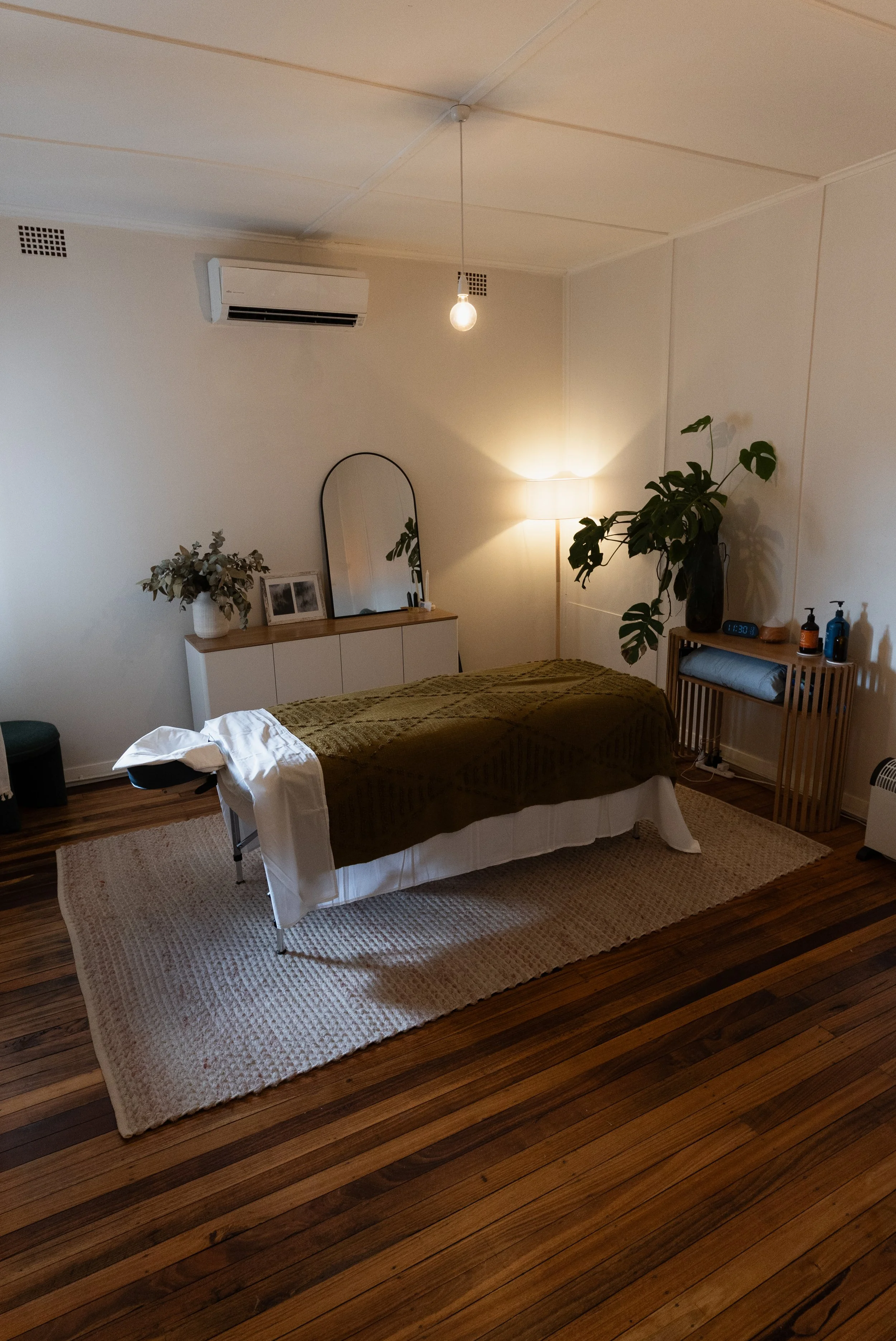 A room with a massage table covered with a green throw, a white sheet hanging off the side, a beige rug underneath, and a wooden floor. There is a side table with a large plant in a dark vase, bottles, and a clock. In the background, a white cabinet 