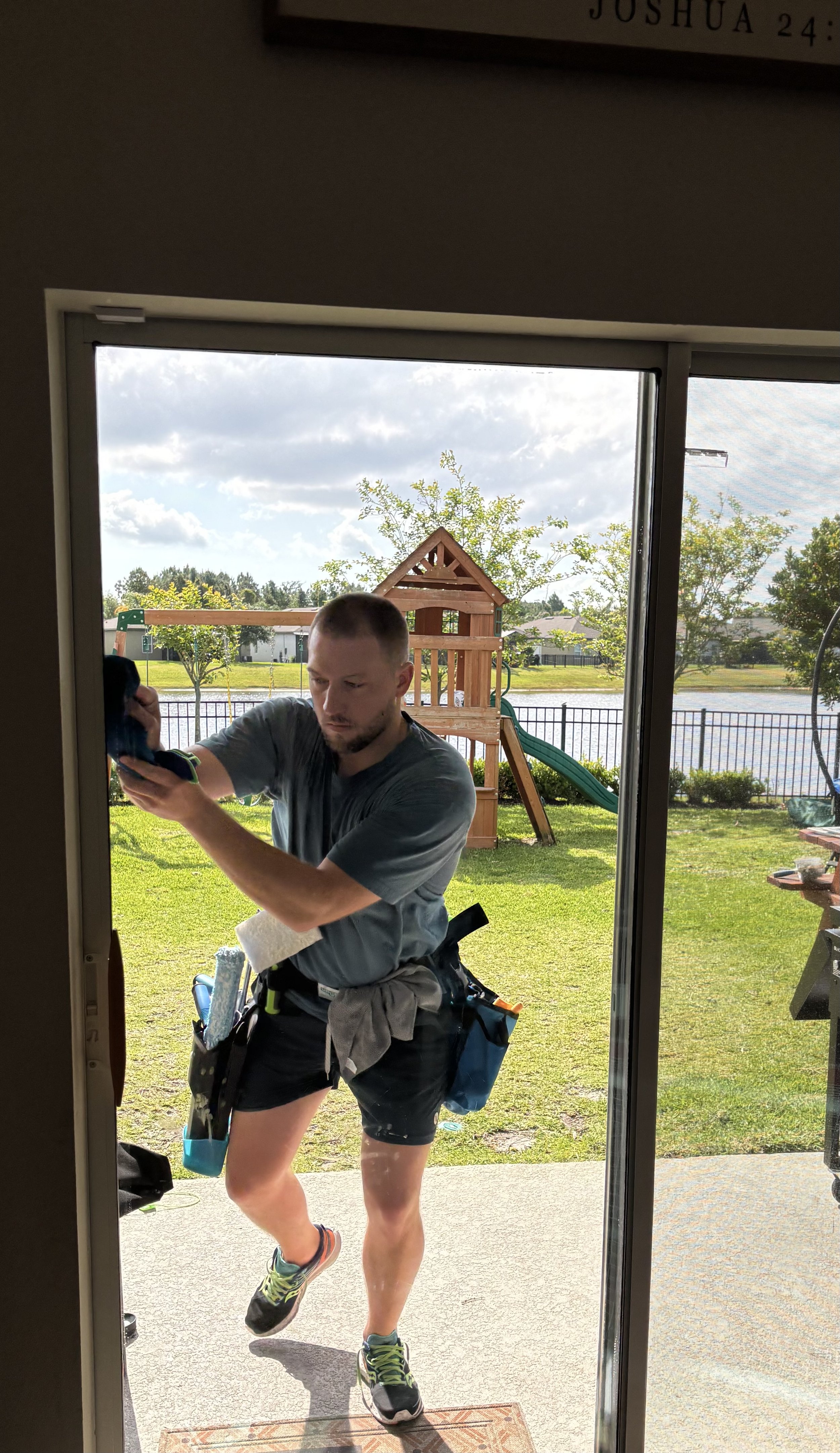 A man is seen through a glass door, holding a phone and appearing to take a selfie or picture. Outside, there is a backyard with a playhouse, trees, a fence, and a lake in the background. The man is wearing a gray shirt, black shorts, running shoes, and has a tool belt around his waist.