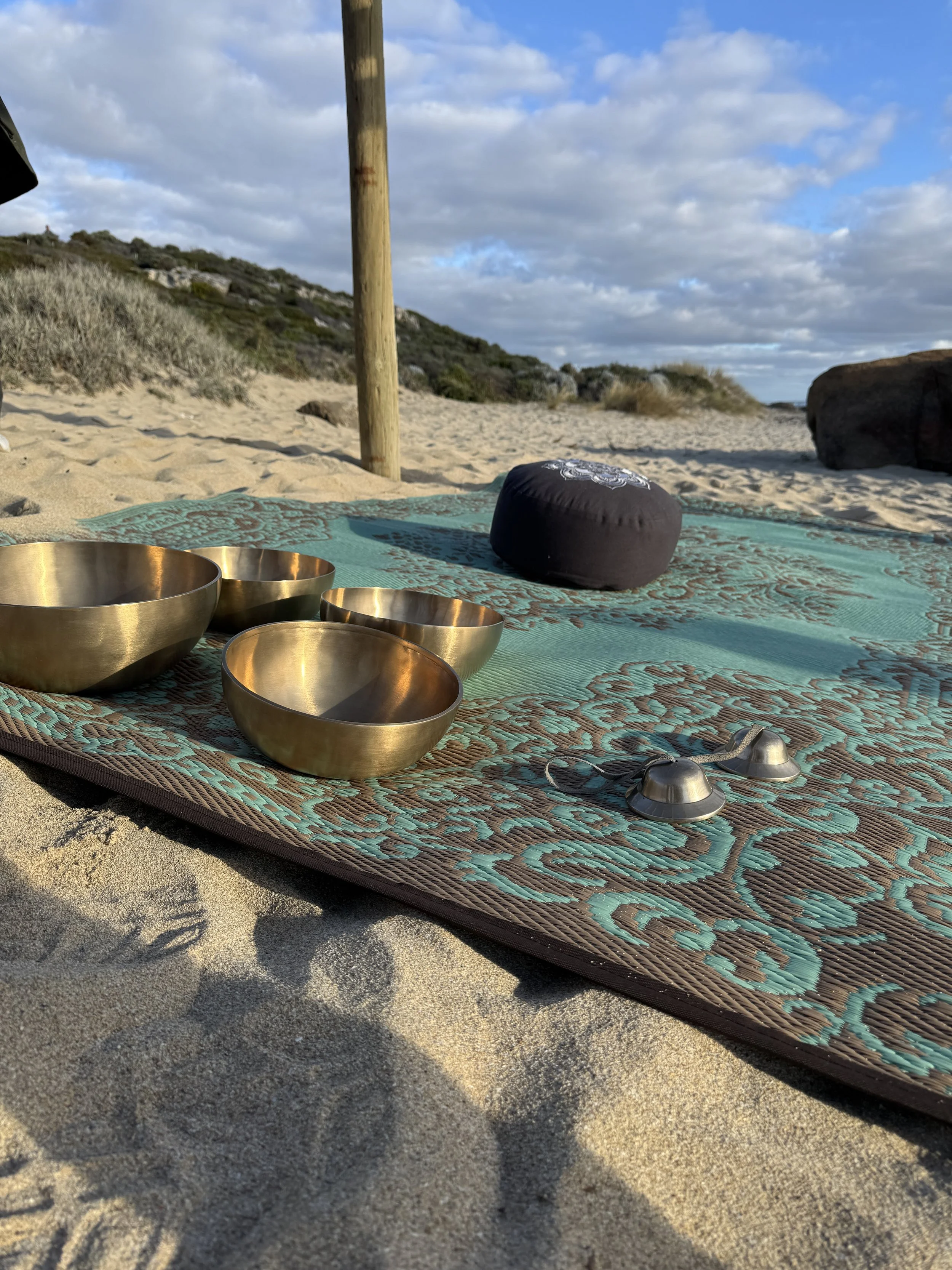 A yoga mat on the sandy beach with singing bowls, a black meditation cushion, and a small bell, with sand dunes, rocks, and a cloudy sky in the background.