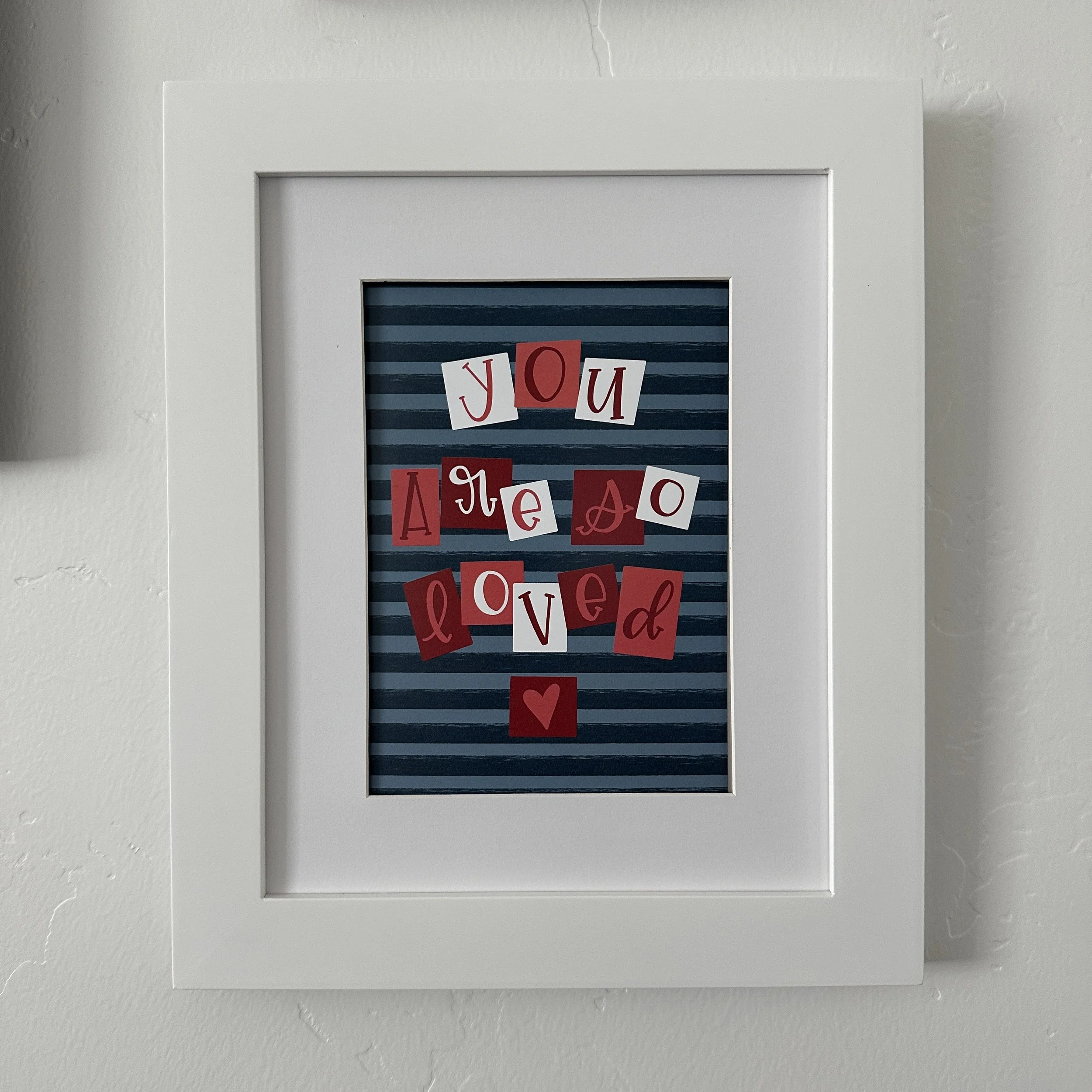 Framed digital print with multicolored letters on a blue striped background spelling 'You are loved' against a white wall.