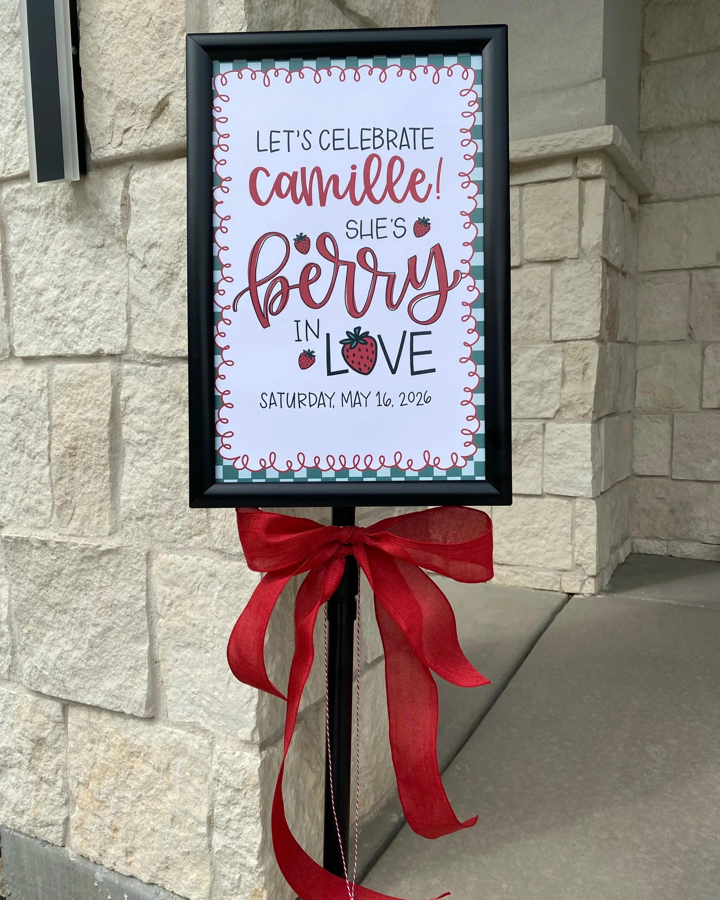 A framed sign with a red ribbon on a sidewalk against a stone wall. The sign reads, 'Let's celebrate Camille! She's berry in love,' with small strawberry illustrations and the date Saturday, May 16, 2026.
