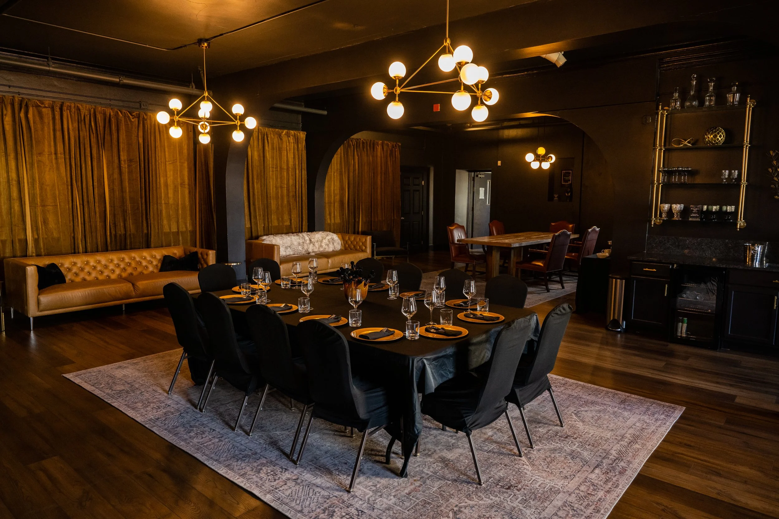 An elegantly decorated dining area with a long table set for a formal meal, surrounded by black chairs. The room features warm lighting with modern chandeliers, gold curtains, leather sofas, and decorative shelves with glassware and ornaments. The space has dark walls and wooden flooring.