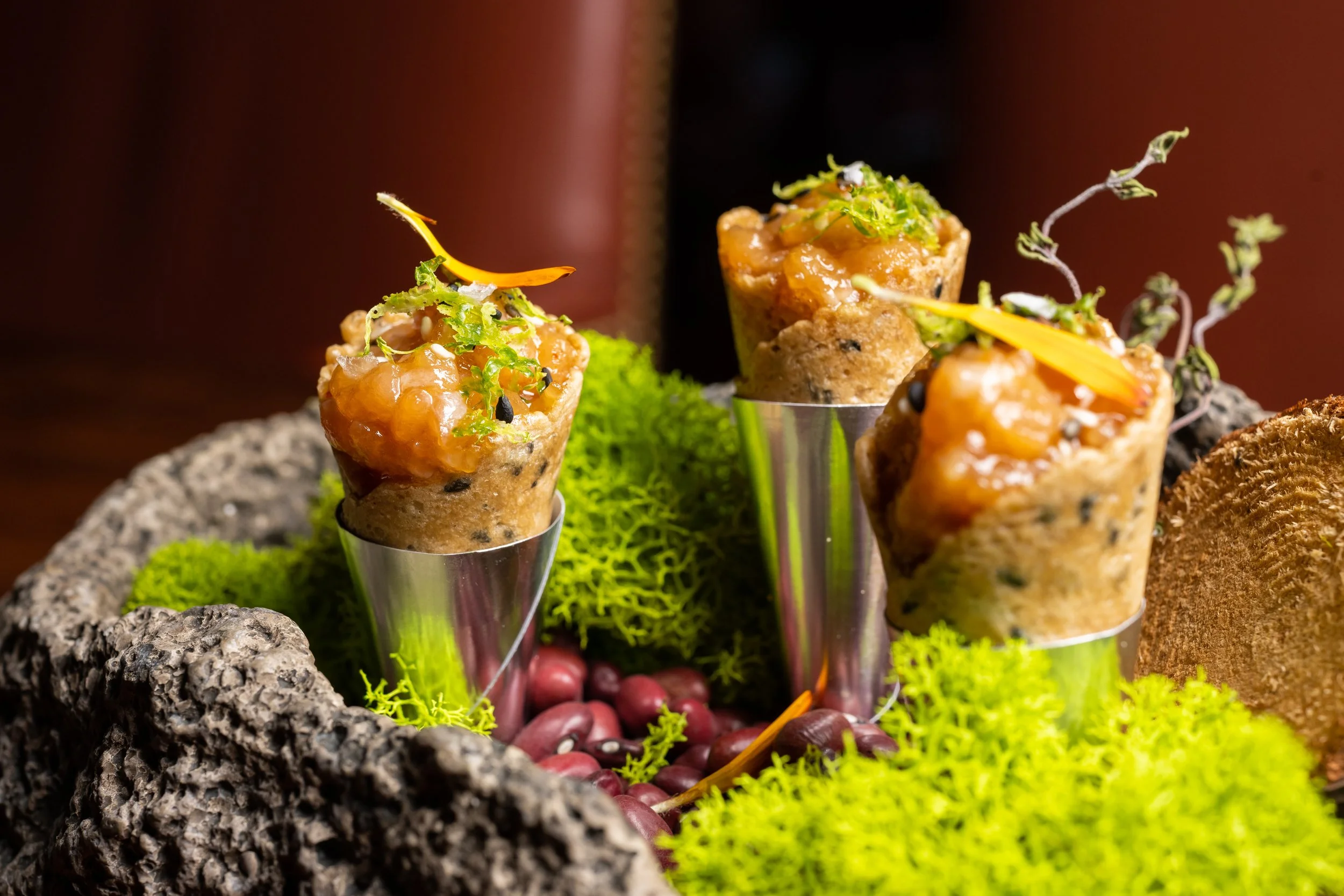 Close-up of gourmet appetizers served in small metal cups, garnished with microgreens and flower petals, on a textured stone platter with green moss and red berries for decoration.