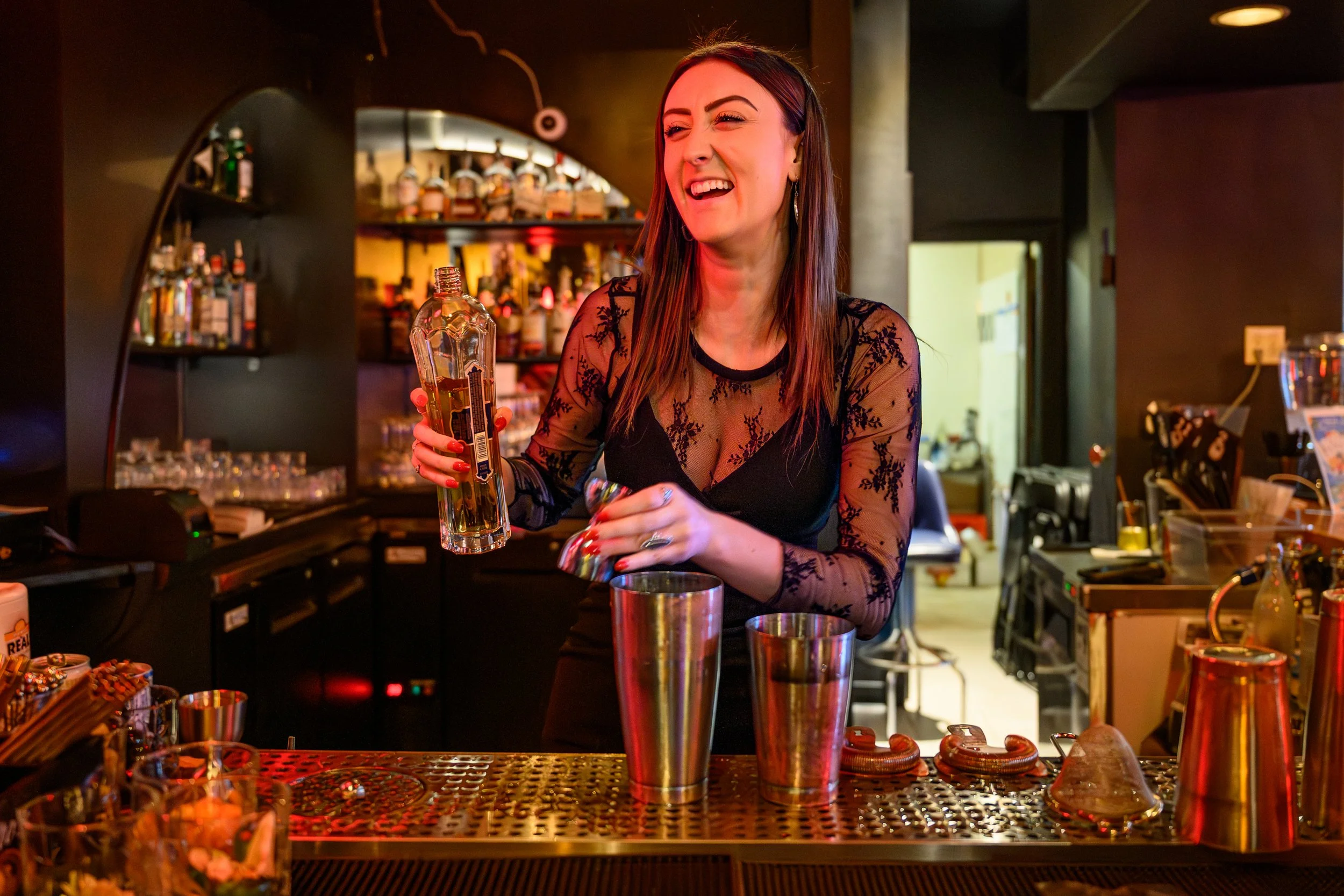 A young woman with long brown hair, wearing a black lace top, is smiling and drinking from a clear bottle behind a bar counter. The bar is decorated with various bottles of alcohol on shelves in the background.