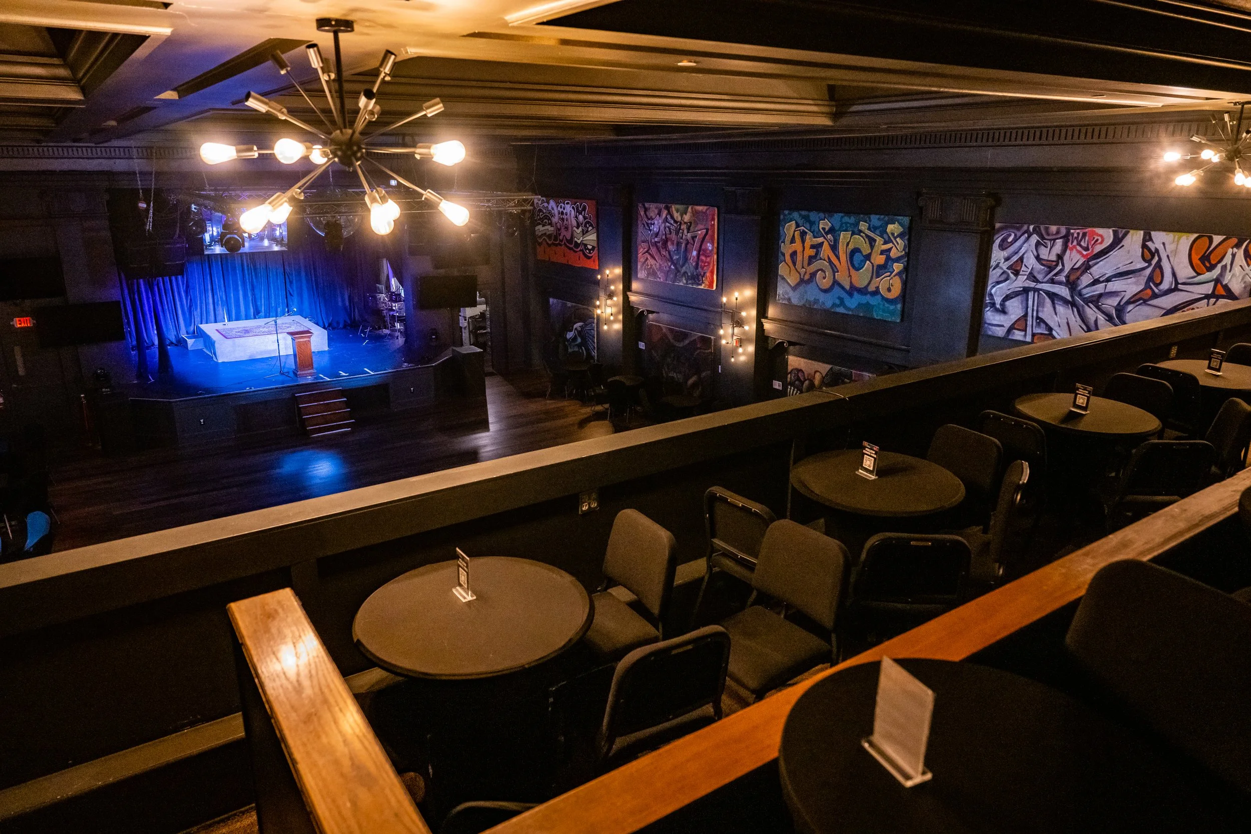 An empty stage with a blue curtain in a dimly lit auditorium, decorated with graffiti-style artwork on the walls, and tables and chairs for the audience in the foreground.