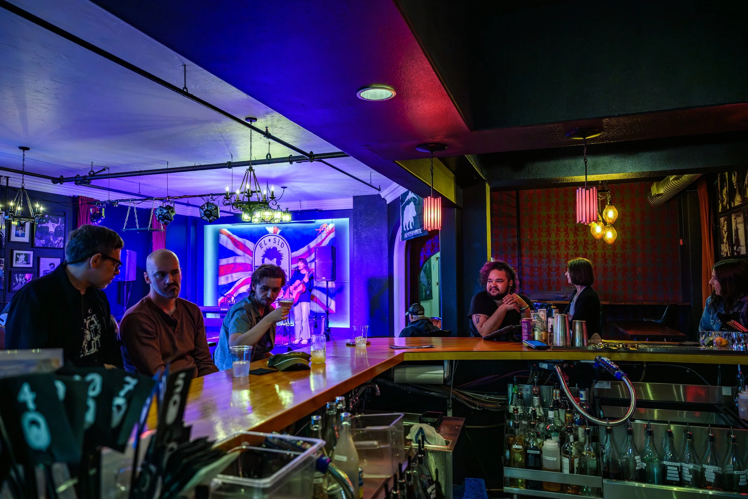 People dining and socializing inside a dimly lit bar or restaurant with colorful neon and stage lighting, featuring a musician performing on a stage with a backdrop, glasses and bottles on the bar counter.