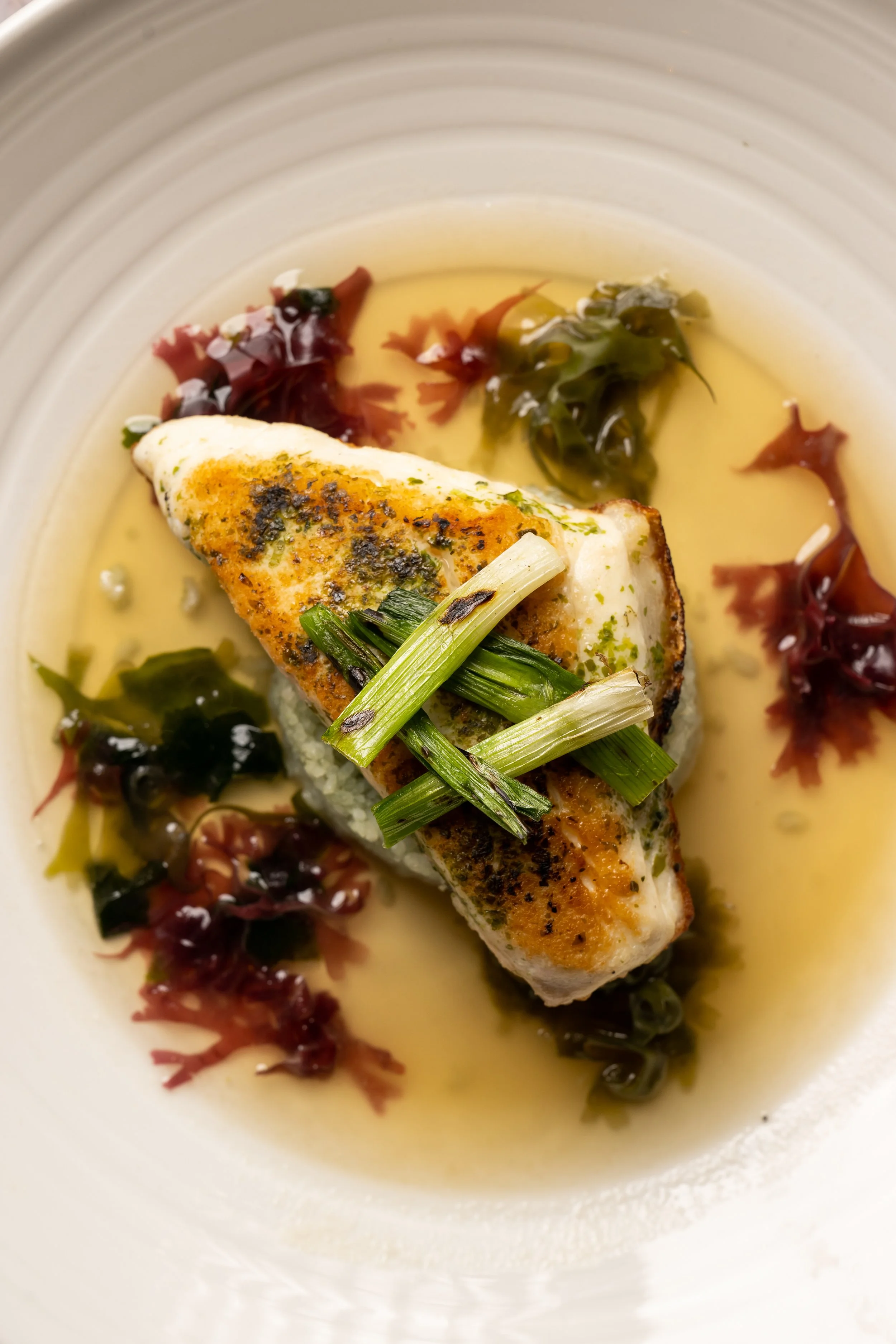 A plate with a cooked fish fillet, garnished with chopped green onions, served over a bed of rice, and accompanied by a sauce with seaweed and sea grapes.