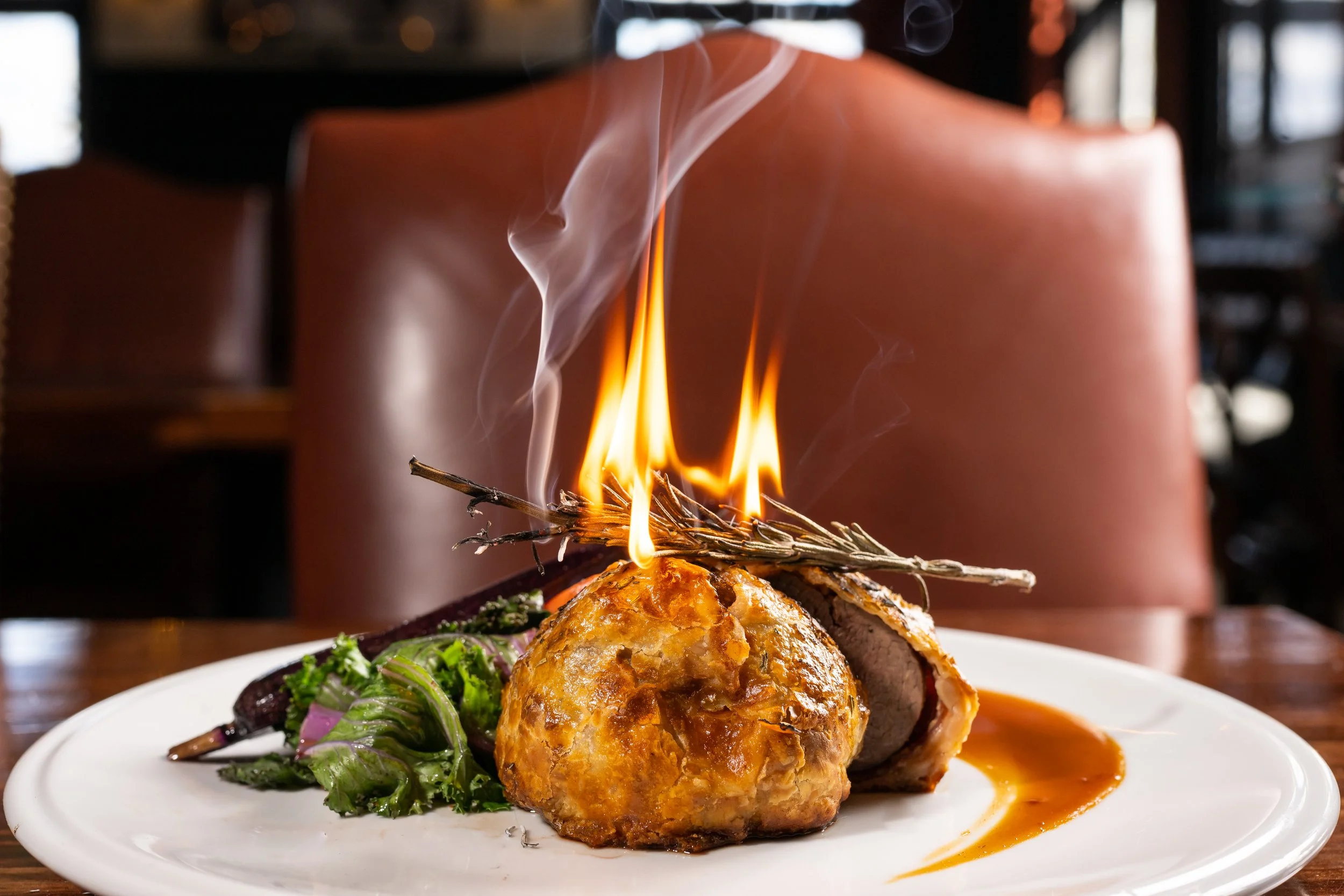 A plate of food with a firework-like flame coming from a dish, accompanied by greens and a sauce, in a restaurant setting.