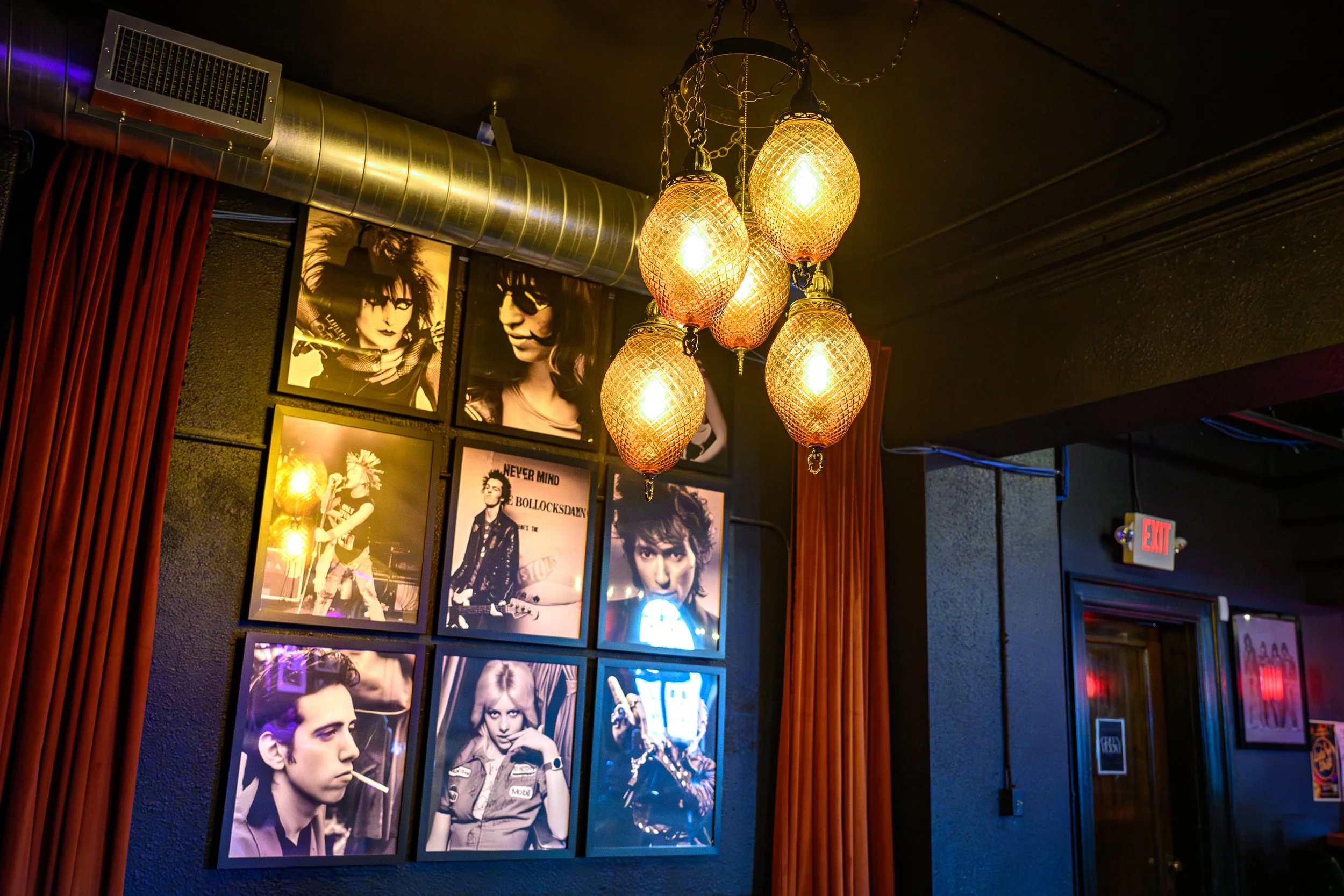 A wall with framed black-and-white photographs of various celebrities and musicians, illuminated by a chandelier with multiple orange-tinted, textured glass lampshades, in a dimly lit room with dark walls, a red curtain, and a visible blue neon sign.