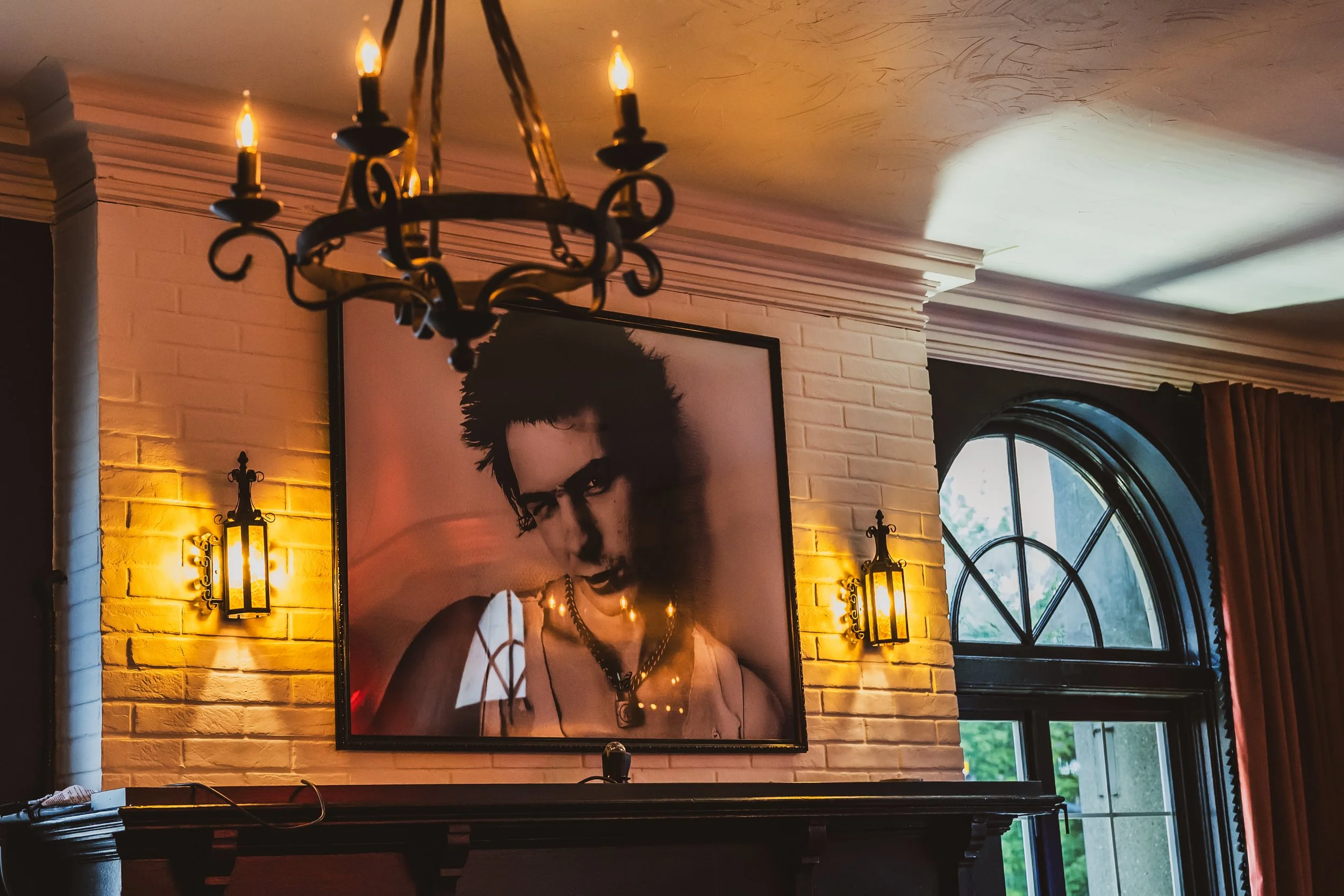 Interior of a room with a white brick wall, featuring a black framed portrait of a woman with short hair and glasses, surrounded by warm yellow wall-mounted lanterns and a black chandelier hanging from the ceiling, with a large arched window showing greenery outside.