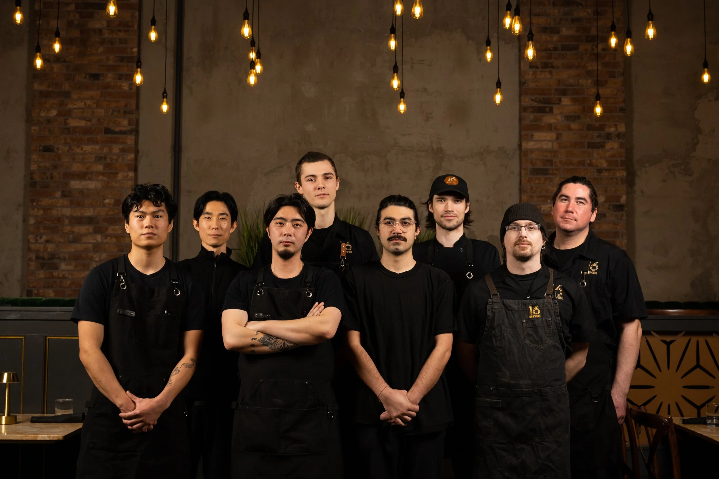 Group of seven diverse male chefs in black uniforms standing in a row in a restaurant with warm lighting and brick walls.