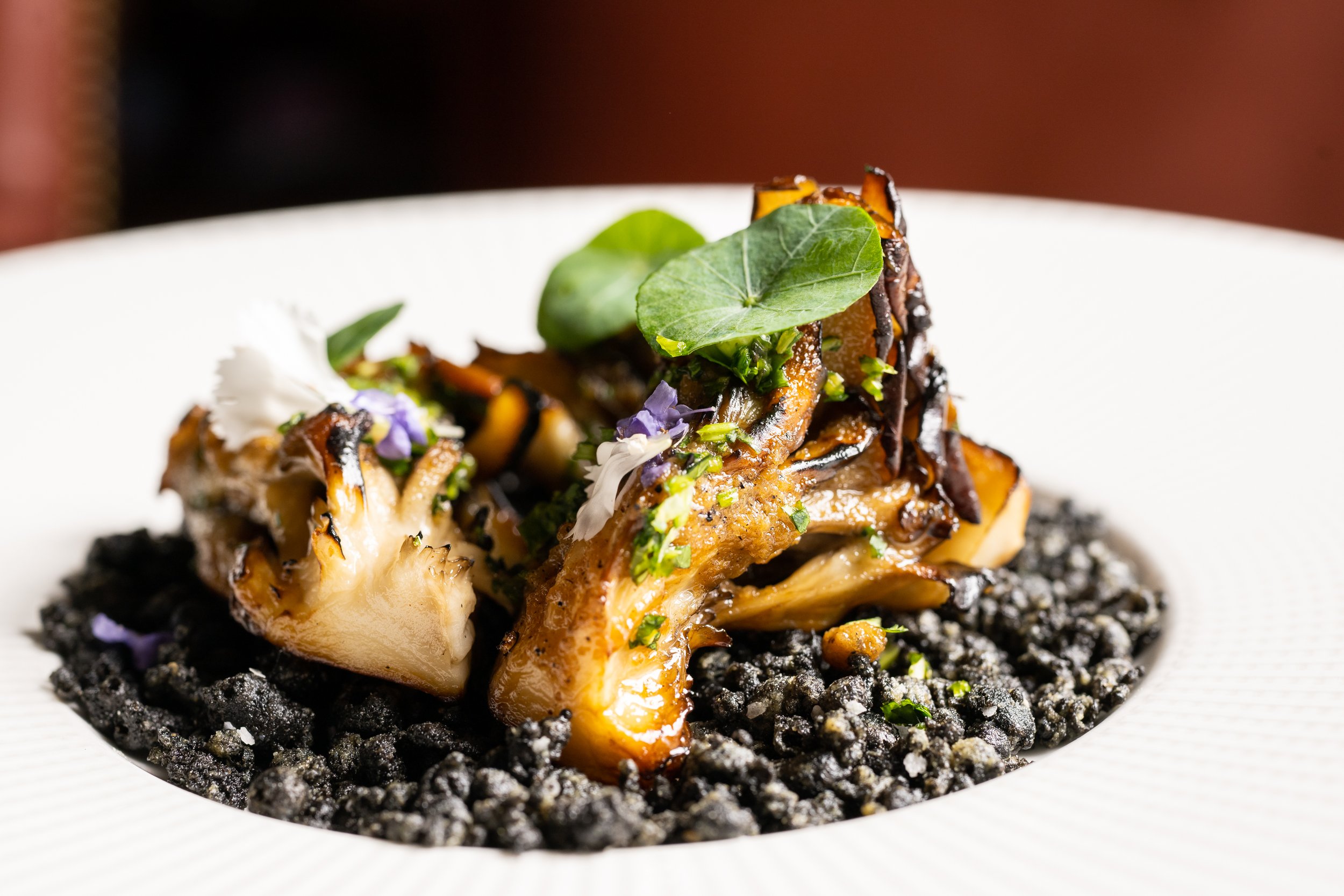 A gourmet dish featuring roasted vegetables and greens on a black crumbly base, garnished with edible flowers and herbs, served on a white plate.