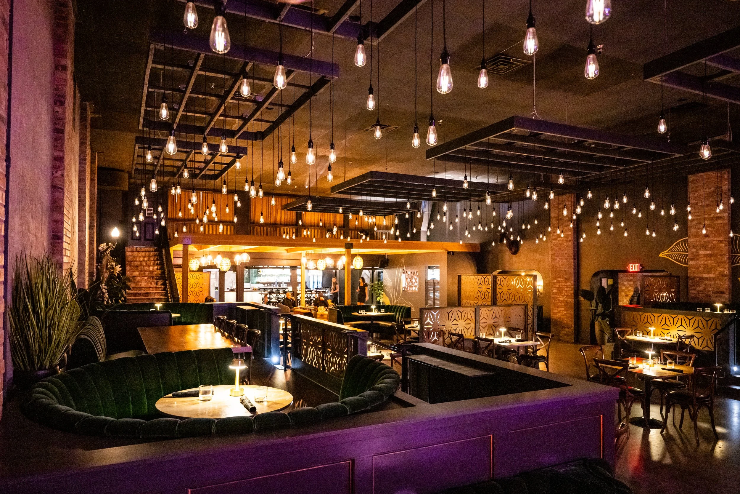 Interior of a dimly lit restaurant or lounge with hanging Edison-style light bulbs, round tables with small lamps, and green velvet booths. There are some patrons and staff in the background near the counter.