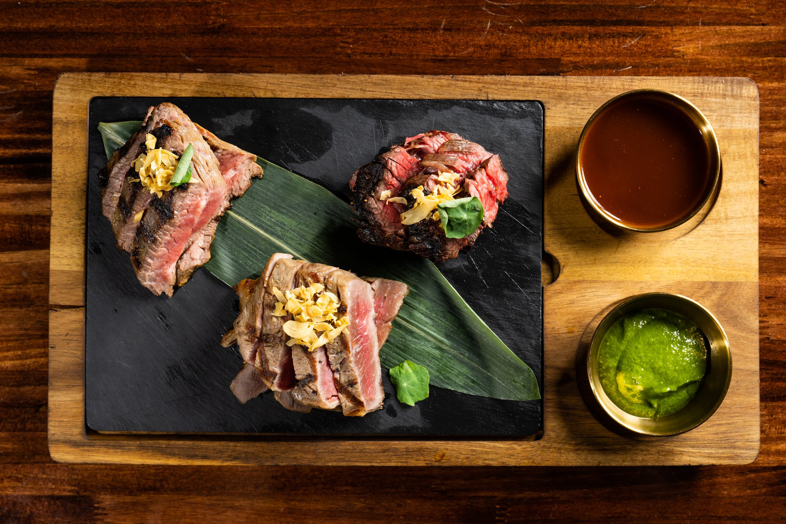 Slices of grilled steak topped with garlic and herbs served on a black slate platter with green leaves, alongside a small bowl of brown sauce and a small bowl of green sauce, on a wooden table.
