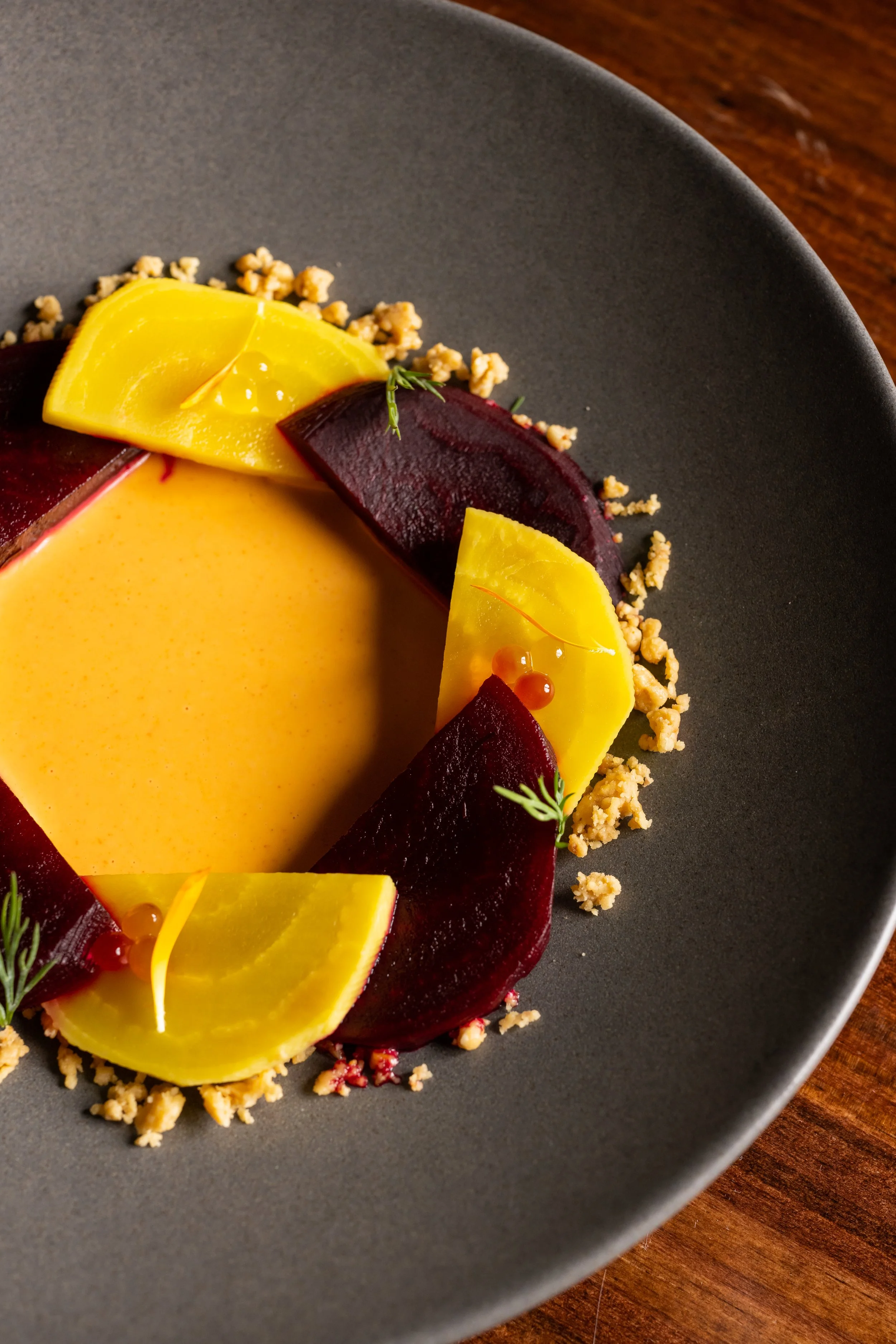Close-up of a plated dessert with yellow and dark red fruit slices, garnished with herbs and crumbs, on a dark gray plate