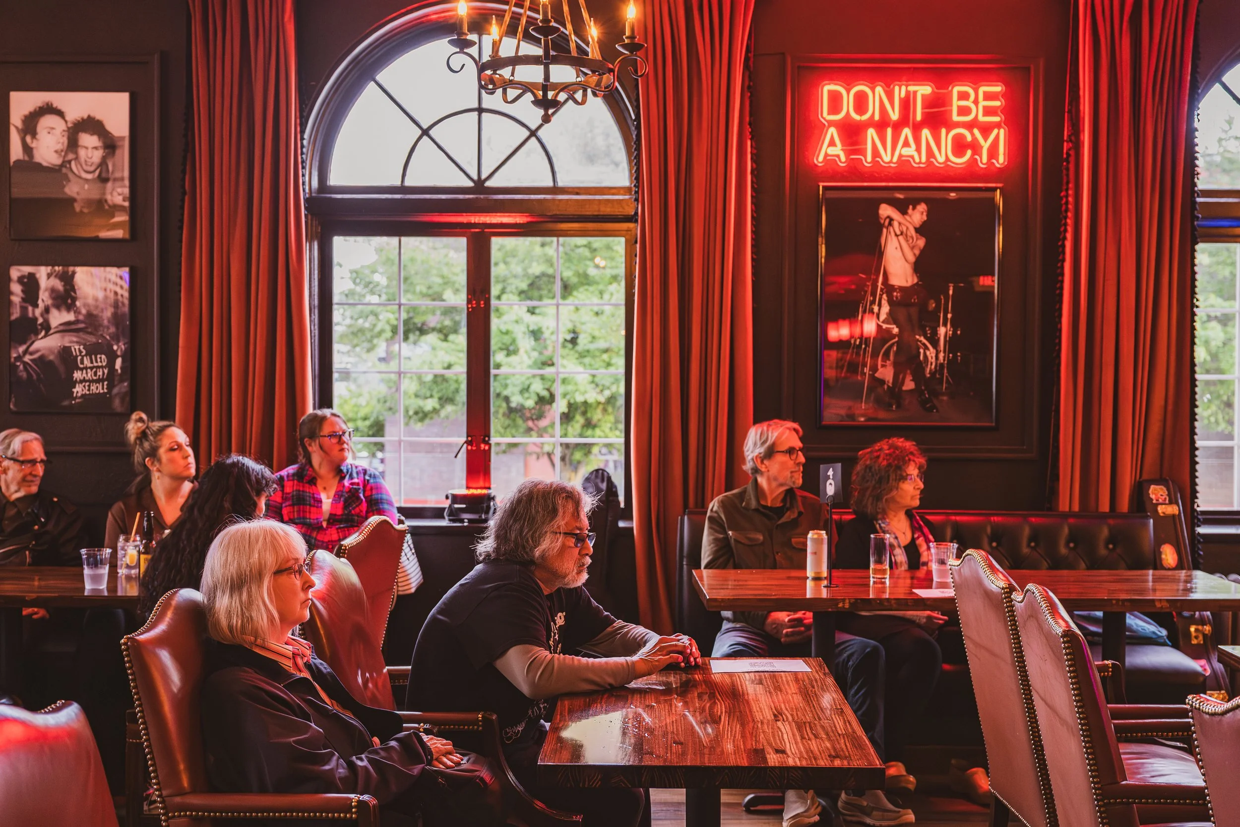 People sitting in a restaurant or cafe, some with drinks in front of them, with large windows, red curtains, and photos on the wall. There is a neon sign that reads 'DON'T BE A NANCY' and a black-and-white portrait of a topless woman.