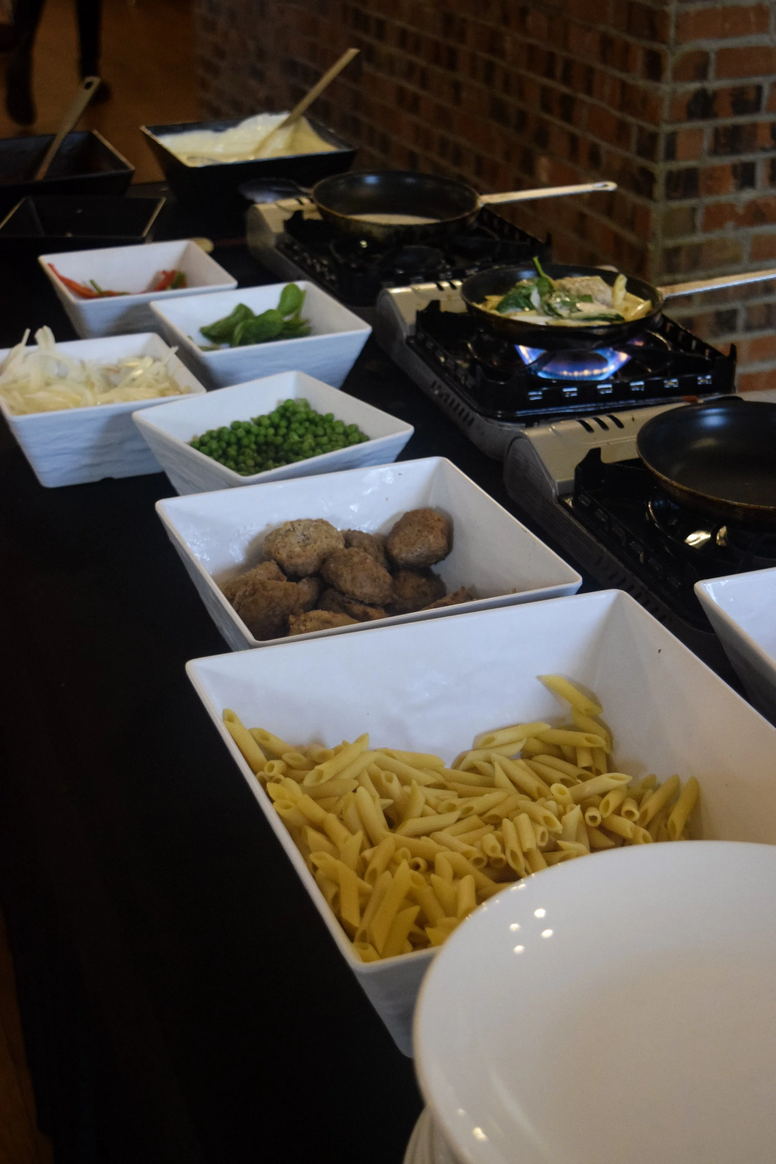 Table with various uncooked ingredients including yellow pasta, fried chicken pieces, green peas, chopped red and green bell peppers, shredded cabbage, and bowls of seasonings or sauces. There are portable stoves with pots and pans, some with cooking