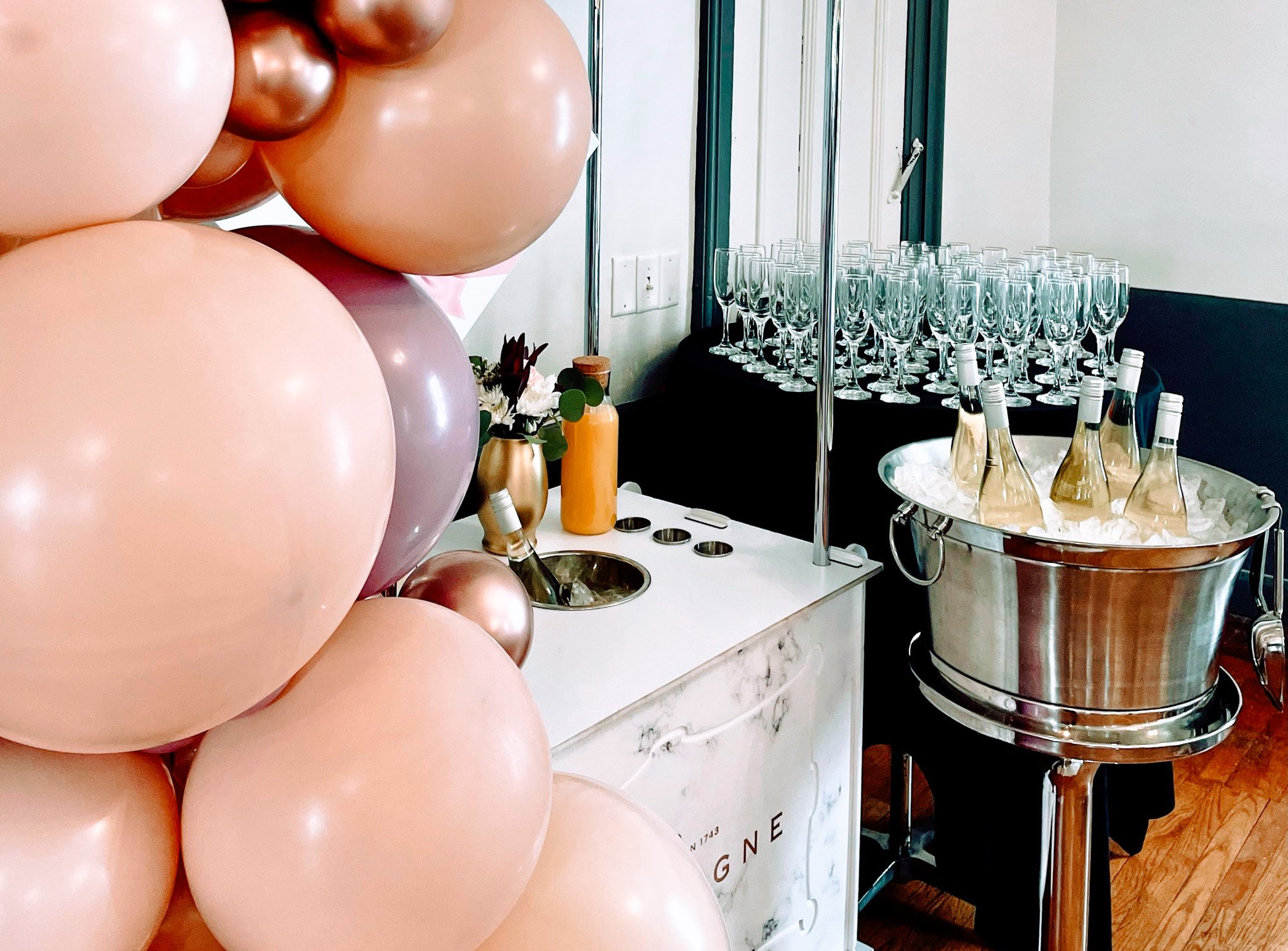A cluster of pink and copper-colored balloons in the foreground, with a table of champagne glasses, bottles, and a floral arrangement in the background, indicating a celebration or event decor.