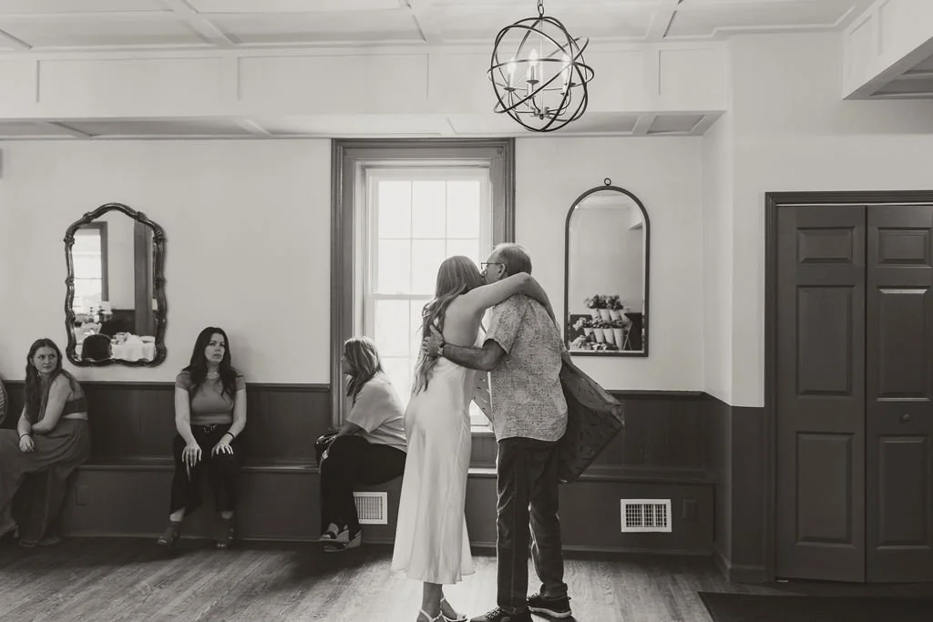 A woman and an older man hugging in a room with other women sitting against the wall, a large window, and decorative mirrors on the wall.